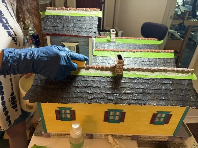 Behind-the-scenes view of the meticulous construction process for the Triple New Orleans Shotgun House Row diorama by Art Illusions by Young. A gloved hand carefully applies mortar to the hand-formed clay ridge tiles along the roof peaks, demonstrati