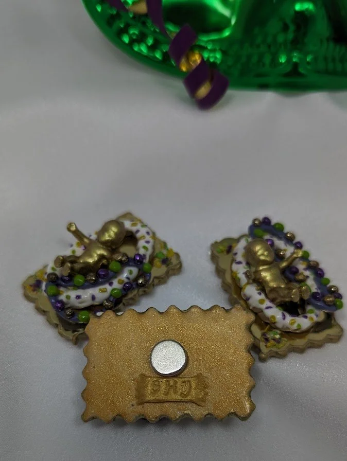 Close-up of three Mardi Gras King Cake Baby magnets/pins by Art Illusions by Young. Intricate hand-sculpted details and painted finishes are visible on the babies, each nestled into a sculpted clay cake on a ornate gold-foiled tray with hand rolled m