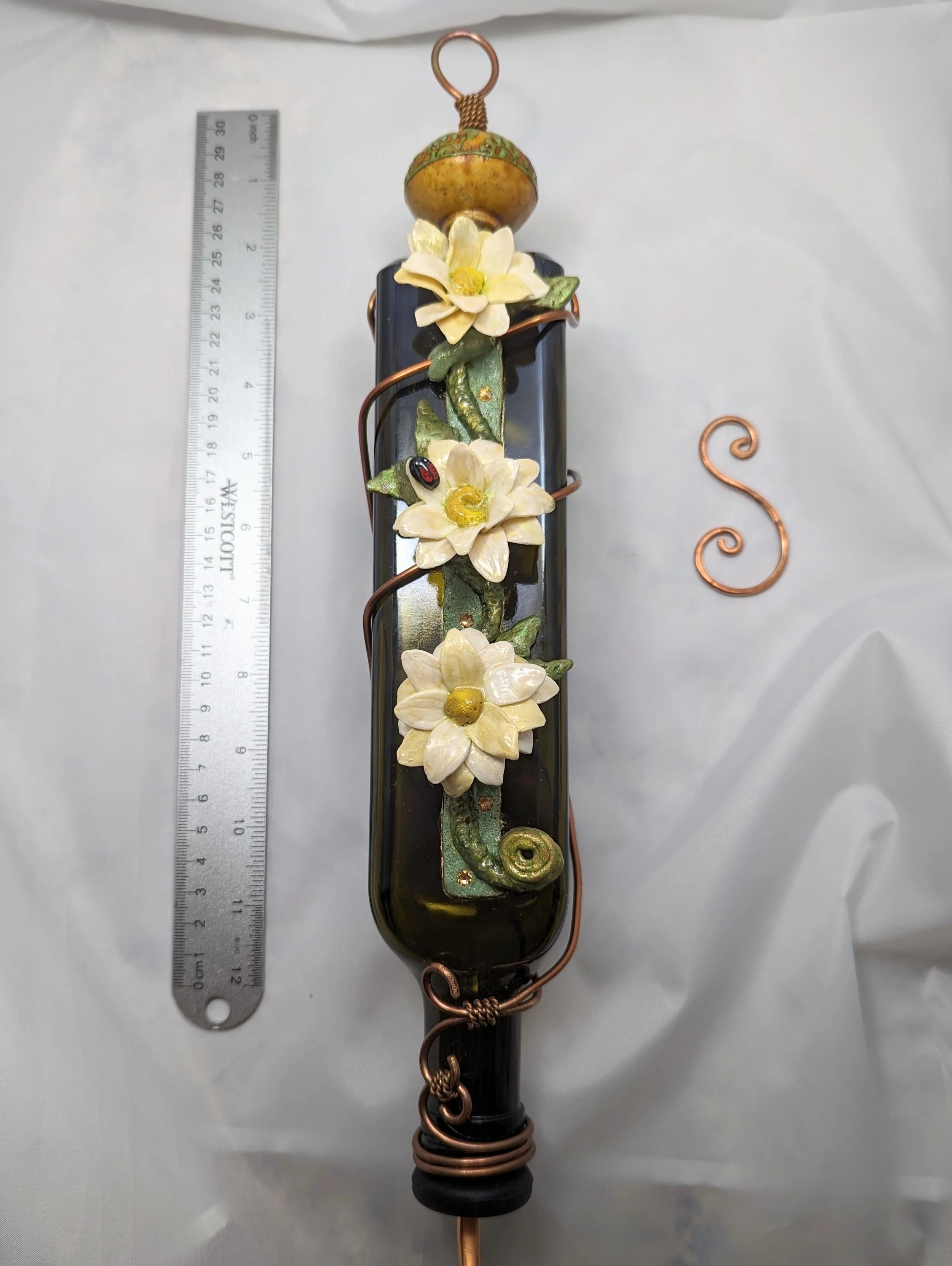 Frontal view of the White Daffodil Elegance Hummingbird Feeder by Art Illusions by Young with ruler for scale. The recycled dark green bottle is adorned with decorative copper wire wrapping and hand-sculpted white daffodils and a tiny ladybug, demons