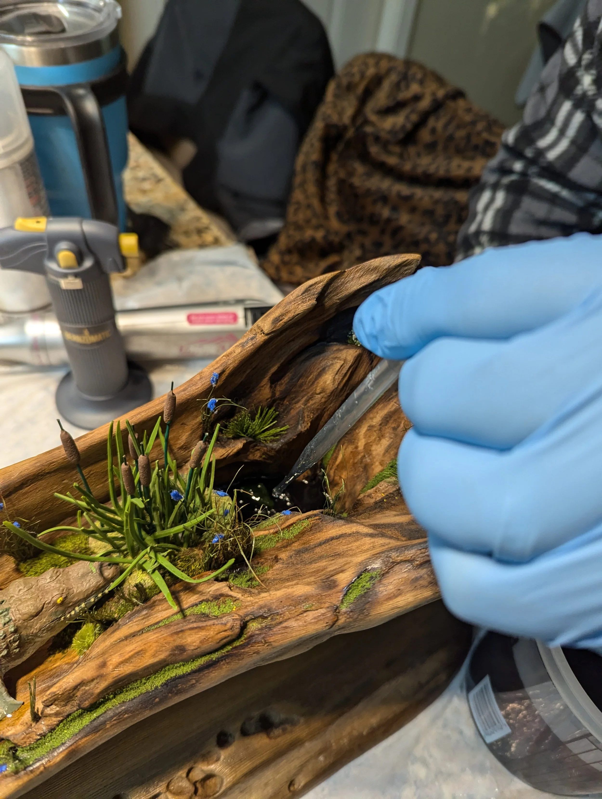 Behind-the-scenes creation of a micro swamp diorama by Art Illusions by Young in Folsom, Louisiana. Gloved hand carefully applying black epoxy water to a textured driftwood base with emerging green plants and small blue accents, showing the detailed 