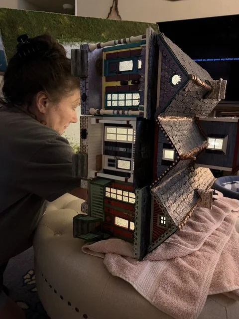 Behind-the-scenes view of artist Sandra Young carefully installing and adjusting the interior lighting in the Triple New Orleans Shotgun House Row diorama at Art Illusions by Young. This intimate photograph captures the focused dedication and precisi
