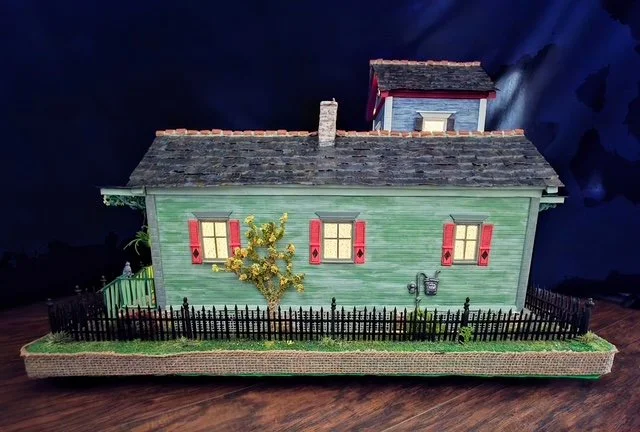 Right-side nighttime view of the handcrafted Triple New Orleans Shotgun House Row diorama by Art Illusions by Young. Warm interior lighting glows through the windows and transoms of the soft sage green shotgun house with striking persimmon orange shu