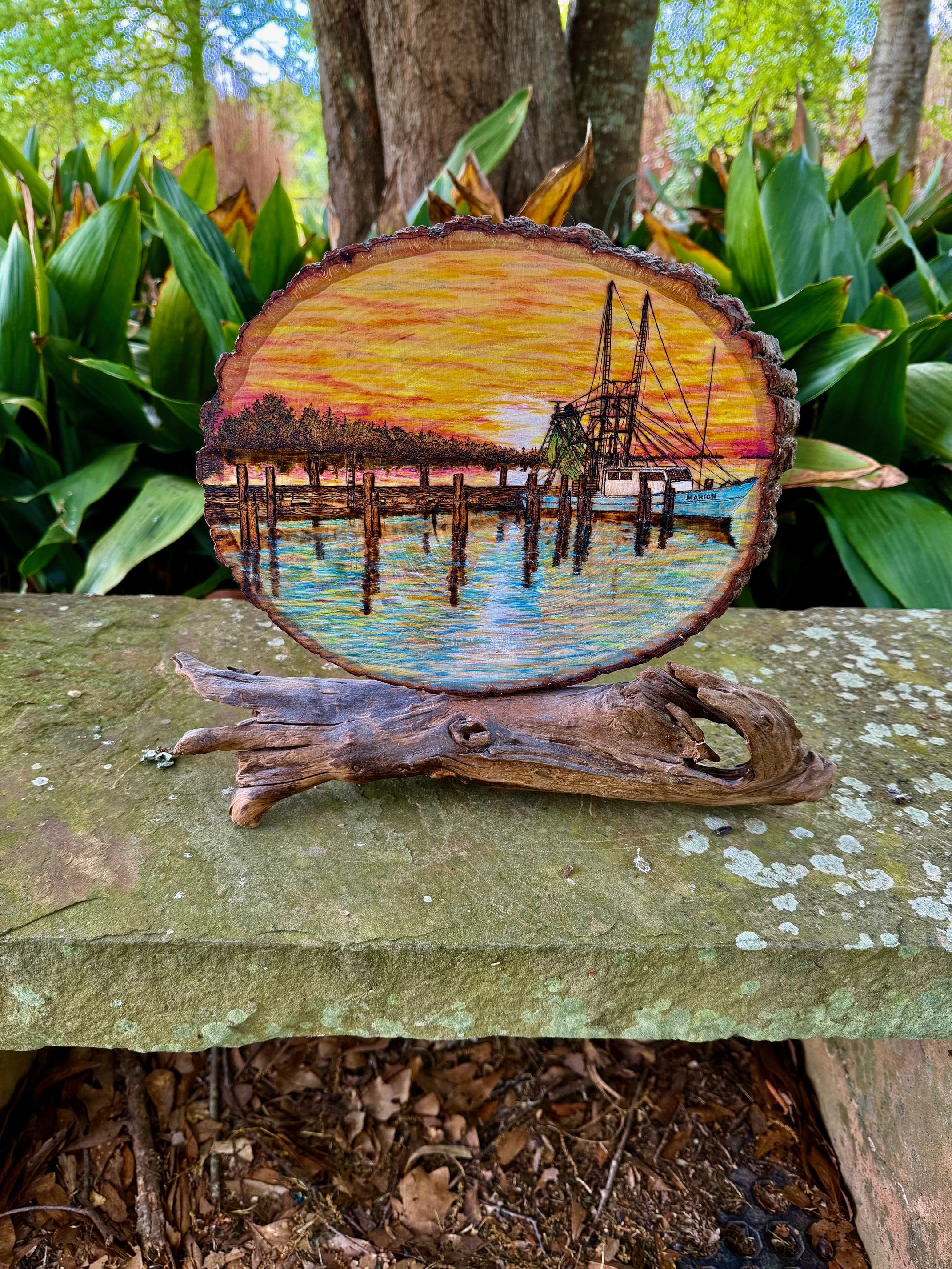 Bucktown Sunset woodburn artwork by Art Illusions by Young displayed among lush plants on stone. The detailed pyrography scene depicts the old fishing village at sunset with warm orange skies reflecting on water, enhanced by hand-applied pigments on 