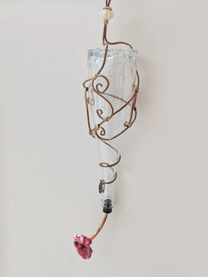 Right side view of the Copper Spiral Grace Hummingbird Feeder by Art Illusions by Young. The clear glass bottle is elegantly encased in decorative copper wire spirals and tendrils, with a single hand-sculpted polymer clay flower enhancing the gracefu