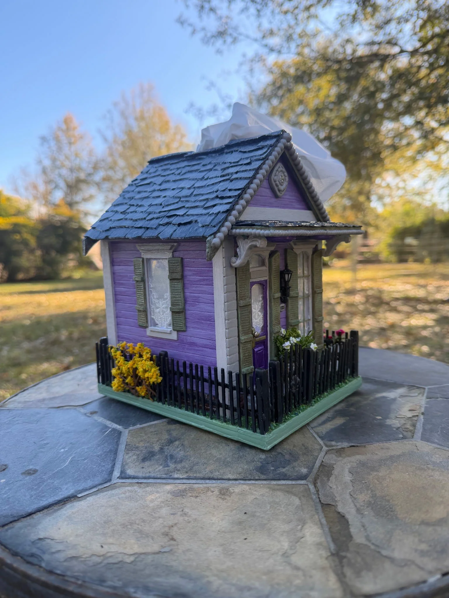 Detailed angled view of the Purple Reign Shotgun House diorama by Art Illusions by Young. Highlights slate rooftop with tissue emerging like a whisper from the Mississippi river, porch lantern, and lush enclosed yard with fabric sheet and flocking ma