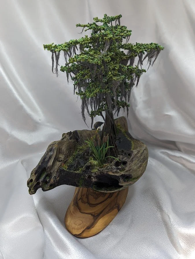 🔴 Sold: 
Moss-Draped Cypress Bonsai Diorama by Art Illusions by Young. A graceful cypress bonsai with long strands of Spanish moss hanging from its branches sits on a natural wood base. The intricate sculpting and textural details evoke the timeless