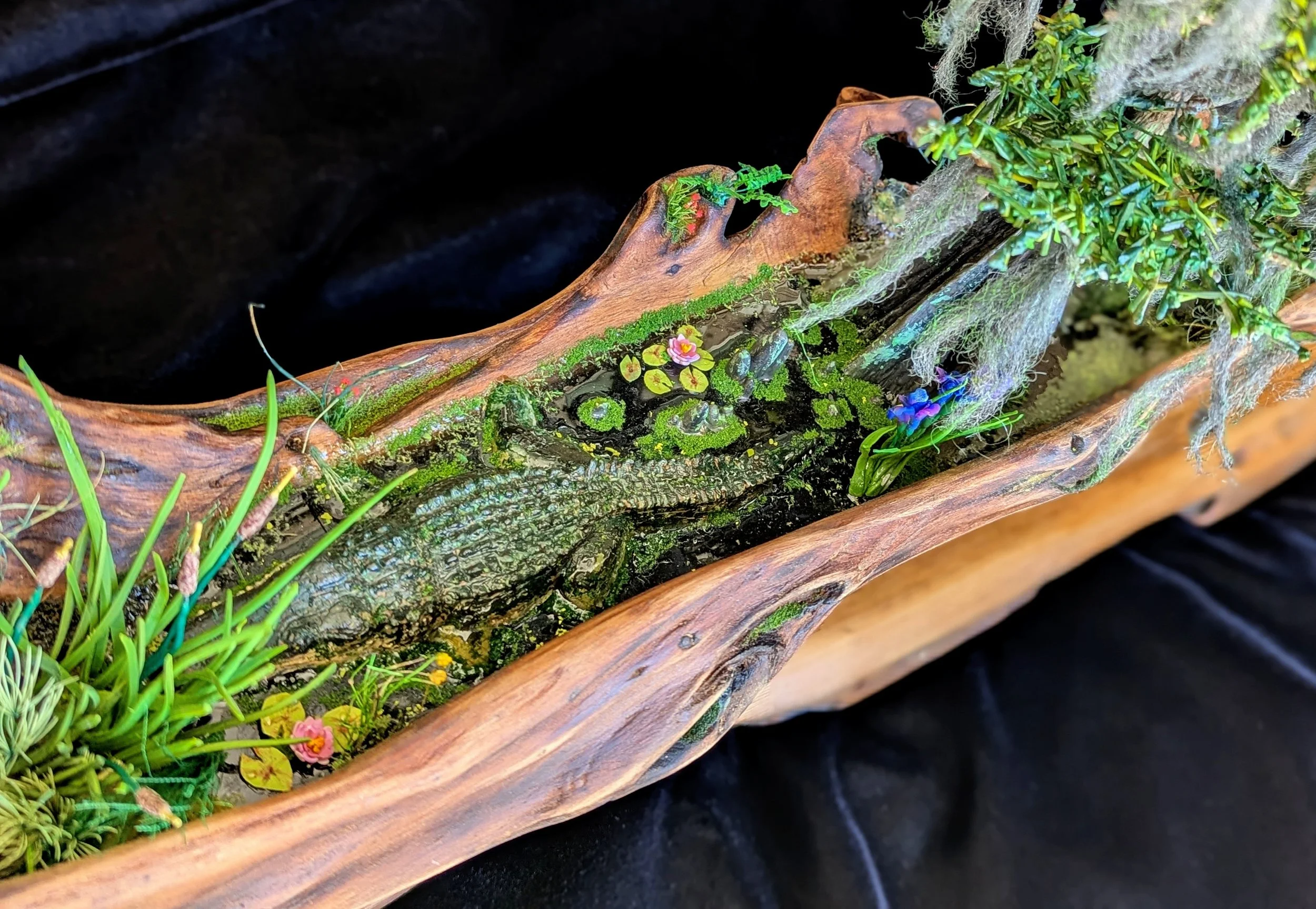 Hand-sculpted miniature alligator with cattails, waterlilies, and tree with Spanish moss on weathered cypress driftwood base. Micro swamp diorama by Art Illusions by Young emphasizing quiet wetland elegance.