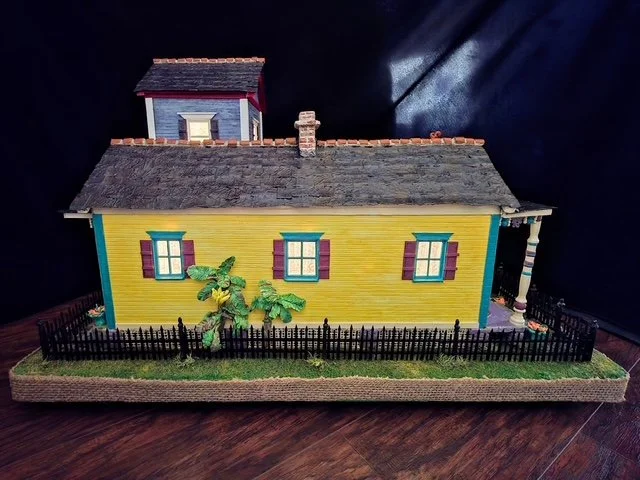 Left-side nighttime view of the handcrafted Triple New Orleans Shotgun House Row diorama by Art Illusions by Young. Warm interior lighting glows softly through the windows, doors, and transoms of the sunny yellow shotgun house with vibrant teal and p