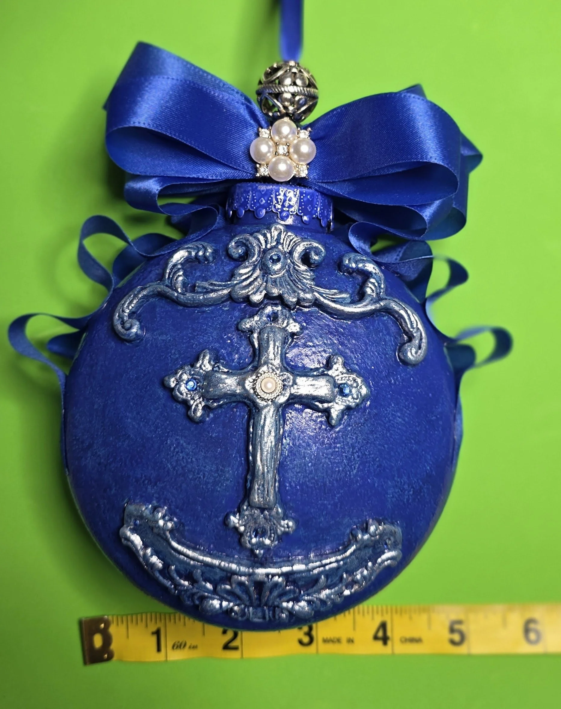 Measured view of the Royal Blue Cross Ornament by Art Illusions by Young showing a width of 5-5.5 inches. The rich cobalt blue surface with metallic sheen and intricate silver cross with raised flourishes are highlighted, while the sparkling ribbon b