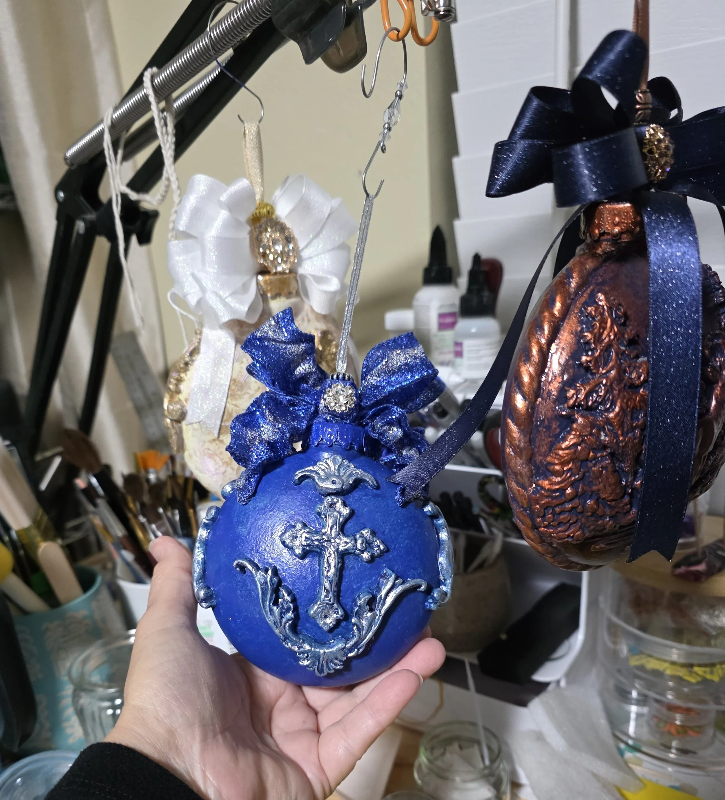 Artist's hand presenting the final stages of the Celestial Blue Cross Ornament by Art Illusions by Young. The luminous cobalt blue surface and intricate silver cross with raised flourishes are visible from multiple angles, while the sparkling royal-b