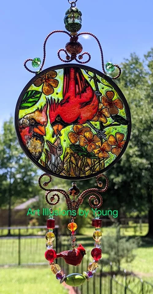 Cardinal Family in Spring’s Painted Light by Art Illusions by Young displayed outdoors. A vibrant male cardinal feeds his hungry fledglings among delicate branches and yellow blossoms. The natural outdoor setting with green foliage enhances the lumin