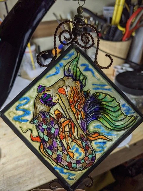 🔴 Sold: 
Mermaid Illusion Glass by Art Illusions by Young. This enchanting diagonal suncatcher depicts a graceful mermaid with flowing hair and tail against a blue aquatic background. Hand-painted details bring fantasy and luminous beauty to the pie