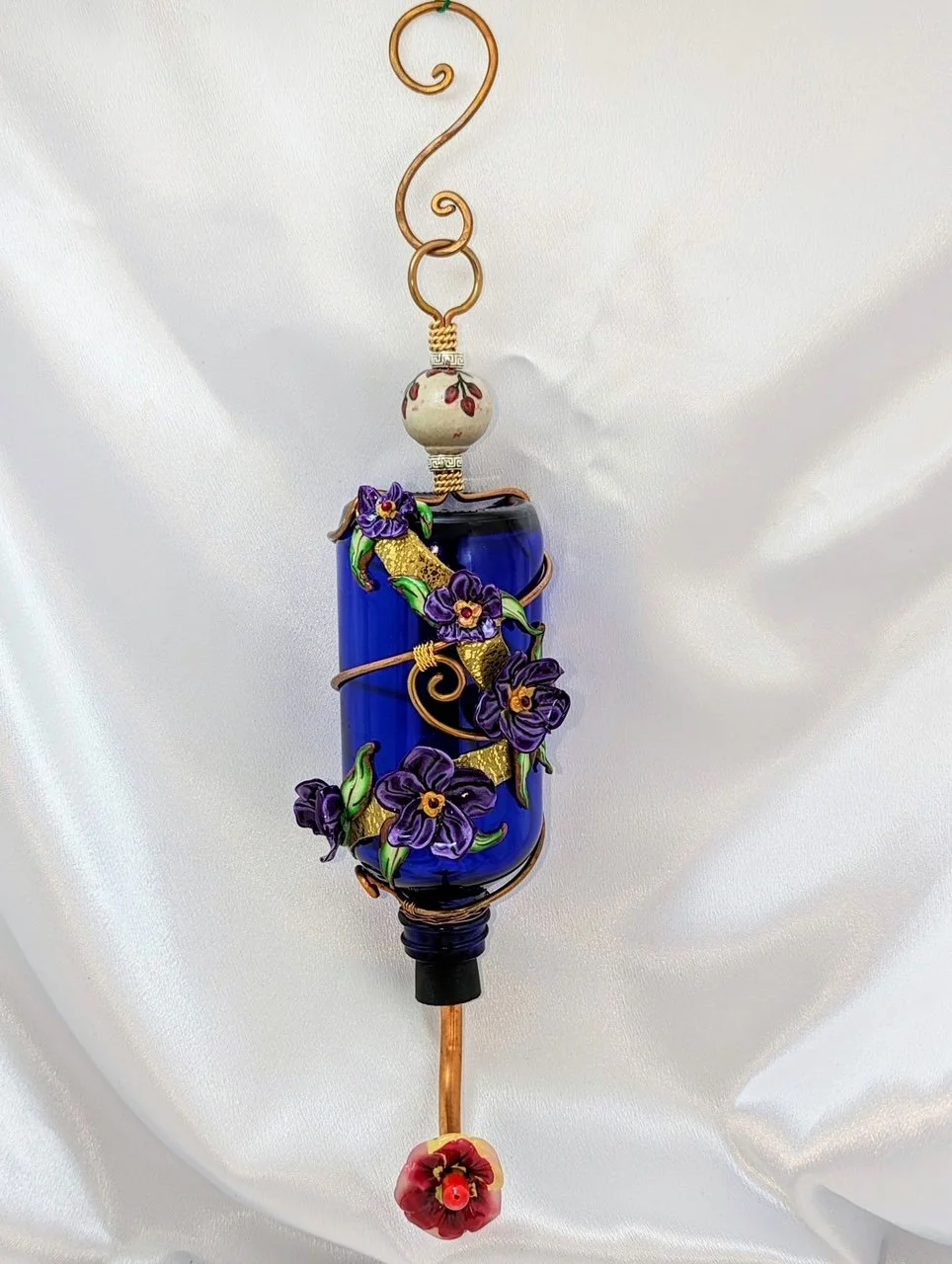 Front view of the Hummingbird Feeder
Purple Pansy Elegance by Art Illusions by Young. The cobalt blue bottle is elegantly encased in decorative copper wire with hand-sculpted purple and violet clay flowers, finished with a secure handmade copper hang