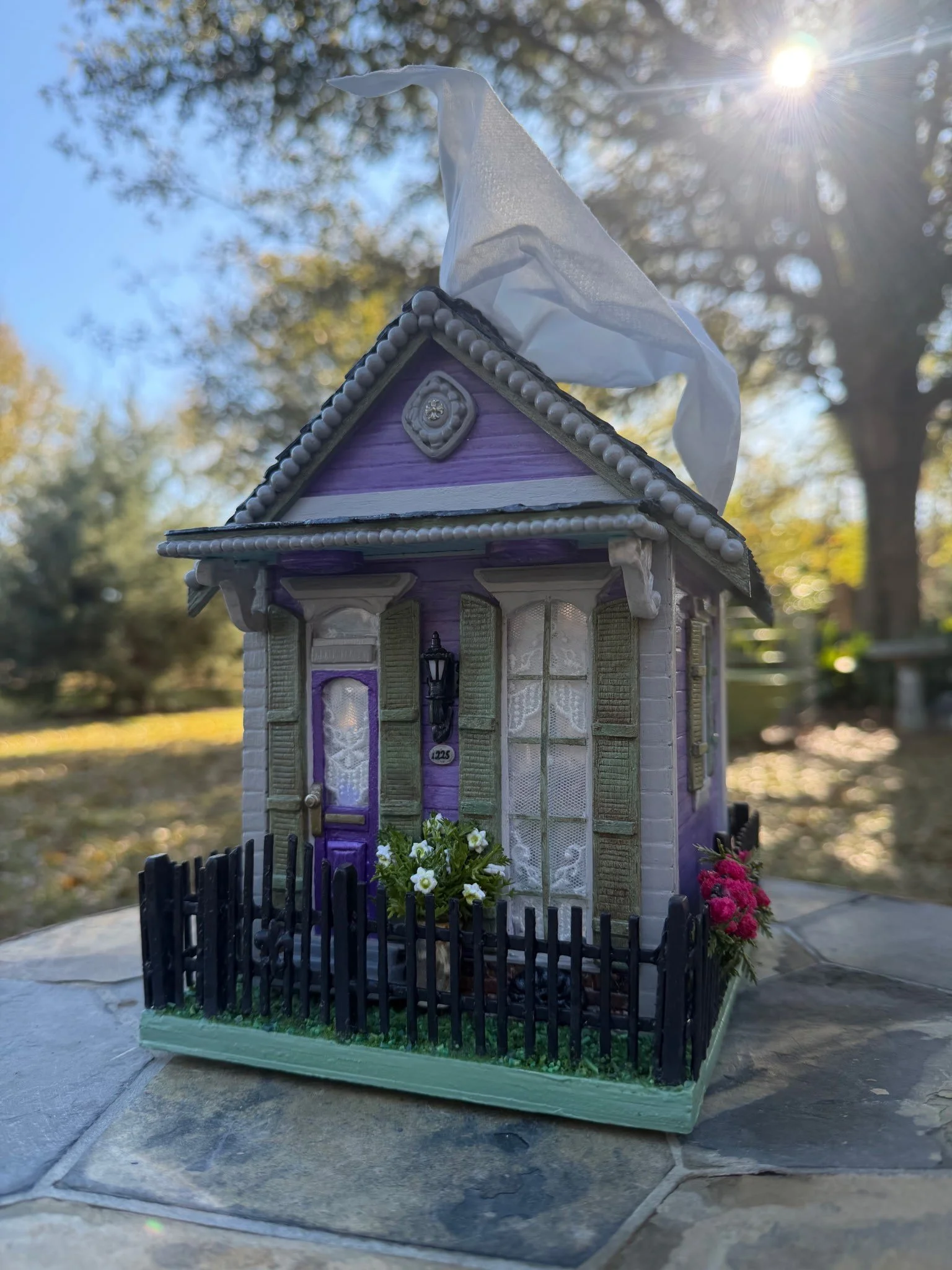 Close front view of the Purple Reign Shotgun House diorama by Art Illusions by Young. Ornate gingerbread trim, reflective lace-curtained doors and windows, golden cross atop the decorative sculpted facade carving, and potted clay gardenia flowers at 
