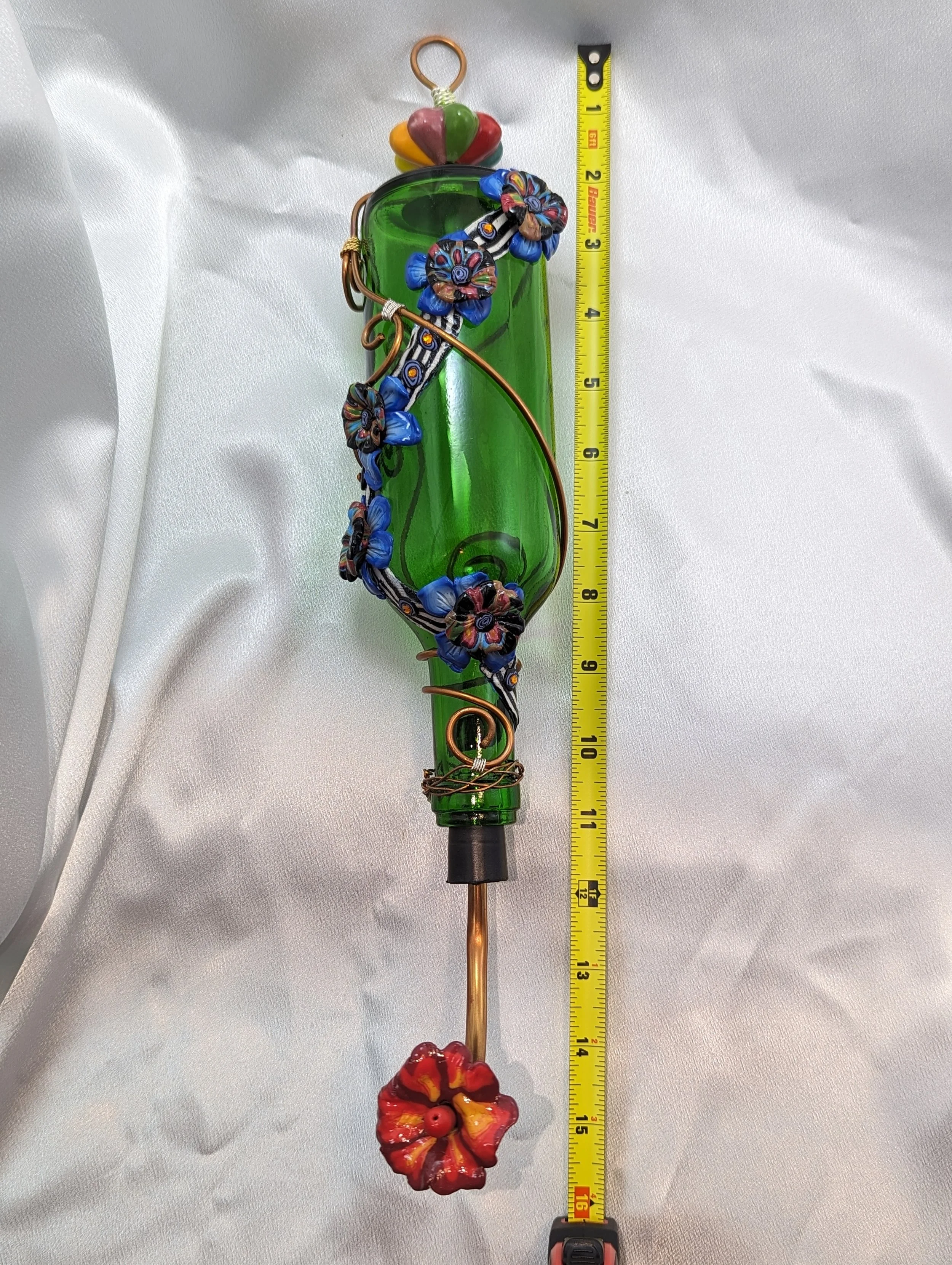 Frontal view with measurement of the Emerald Garden Bloom Hummingbird Feeder by Art Illusions by Young with ruler for scale. The recycled emerald green glass bottle is adorned with intricate copper wire vines and hand-sculpted blue and multicolored p