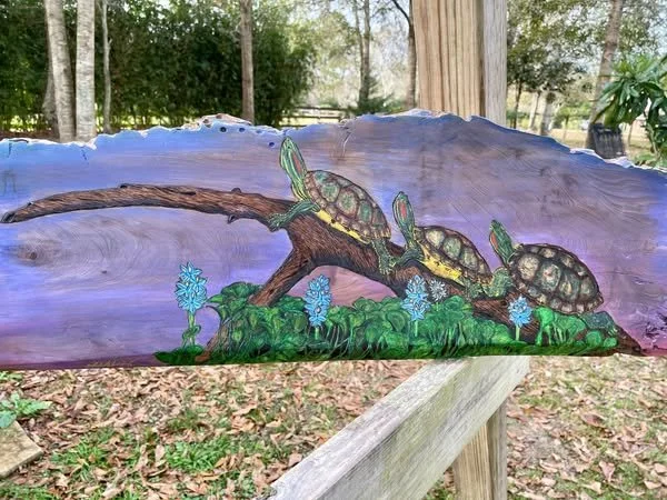 Red-Eared Slider Turtles pyrography on live-edge sinker cypress by Art Illusions by Young from Folsom, Louisiana. Three red-eared slider turtles bask on a fallen log above a reflective water edge, surrounded by blooming blue irises and a wooded bayou