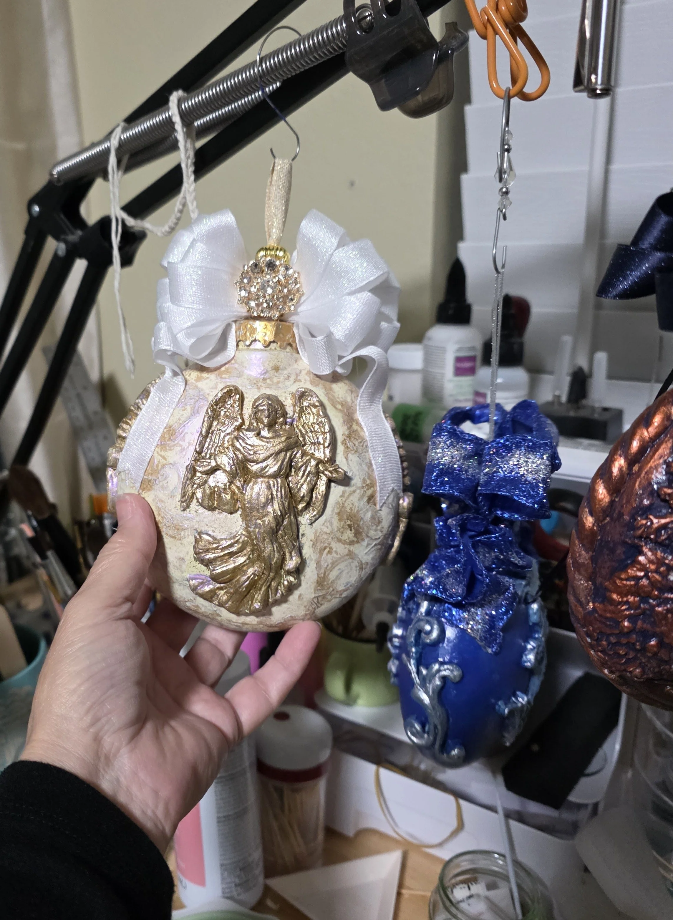A hand-sculpted and molded Guardian Angel Ornament by Art Illusions by Young is held in a studio setting, shown prior to final completion details. The detailed angel figure, rendered in luminous iridescent pearl, blush, and golden tones, features out