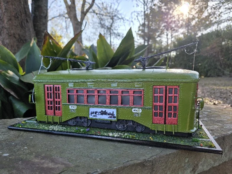 Handcrafted New Orleans St. Charles Avenue streetcar diorama by Art Illusions by Young. Olive green body with crimson windows and doors, authentic tracks, grassy base with wildflowers, and glowing interior LED lights. Functional tissue box cover mixe