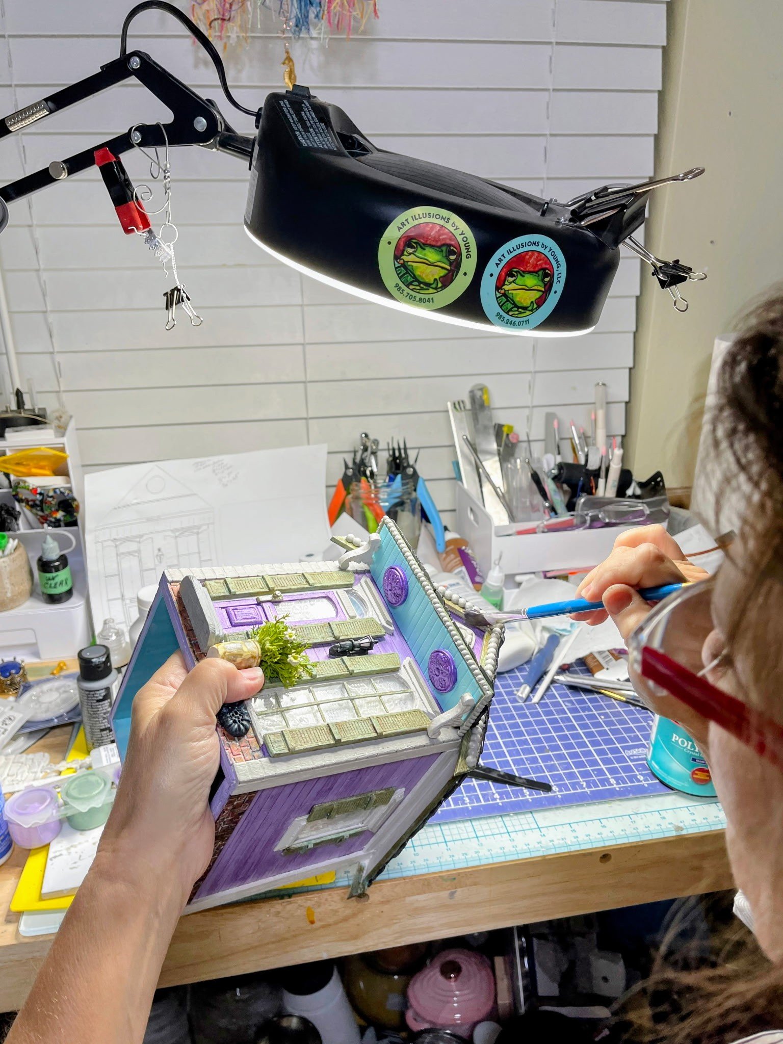 Artist’s hands carefully assembling and detailing the Purple Reign Shotgun House tissue box cover diorama. Shows work on the porch details, and polymer clay elements. Studio craftsmanship by Art Illusions by Young bringing historic New Orleans archit