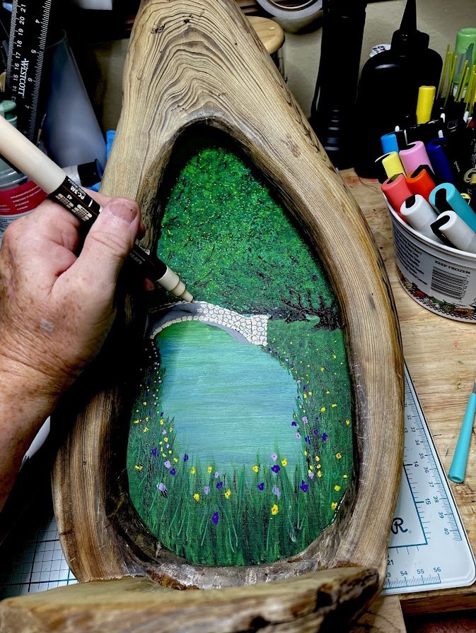 Close-up of artist’s hand carefully painting and detailing the miniature pond and bridge inside the Enchanted City Park Serenity diorama. Studio craftsmanship by Art Illusions by Young.