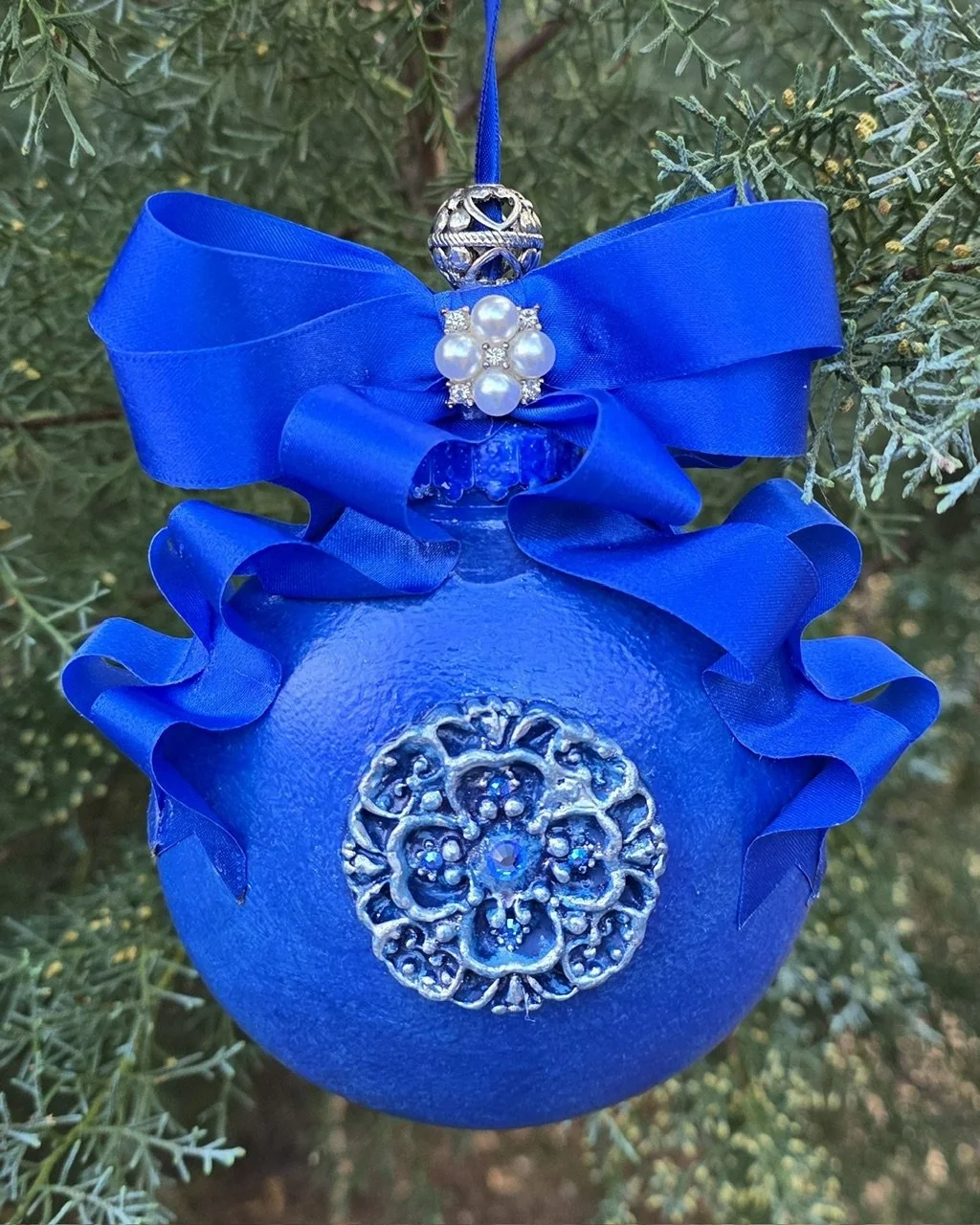 Rear close-up view of the Royal Blue Cross Ornament by Art Illusions by Young. The cobalt blue orb and ornate silver cross with filigree detailing are highlighted against green foliage, crowned by a royal-blue ribbon bow. This compact piece brings qu