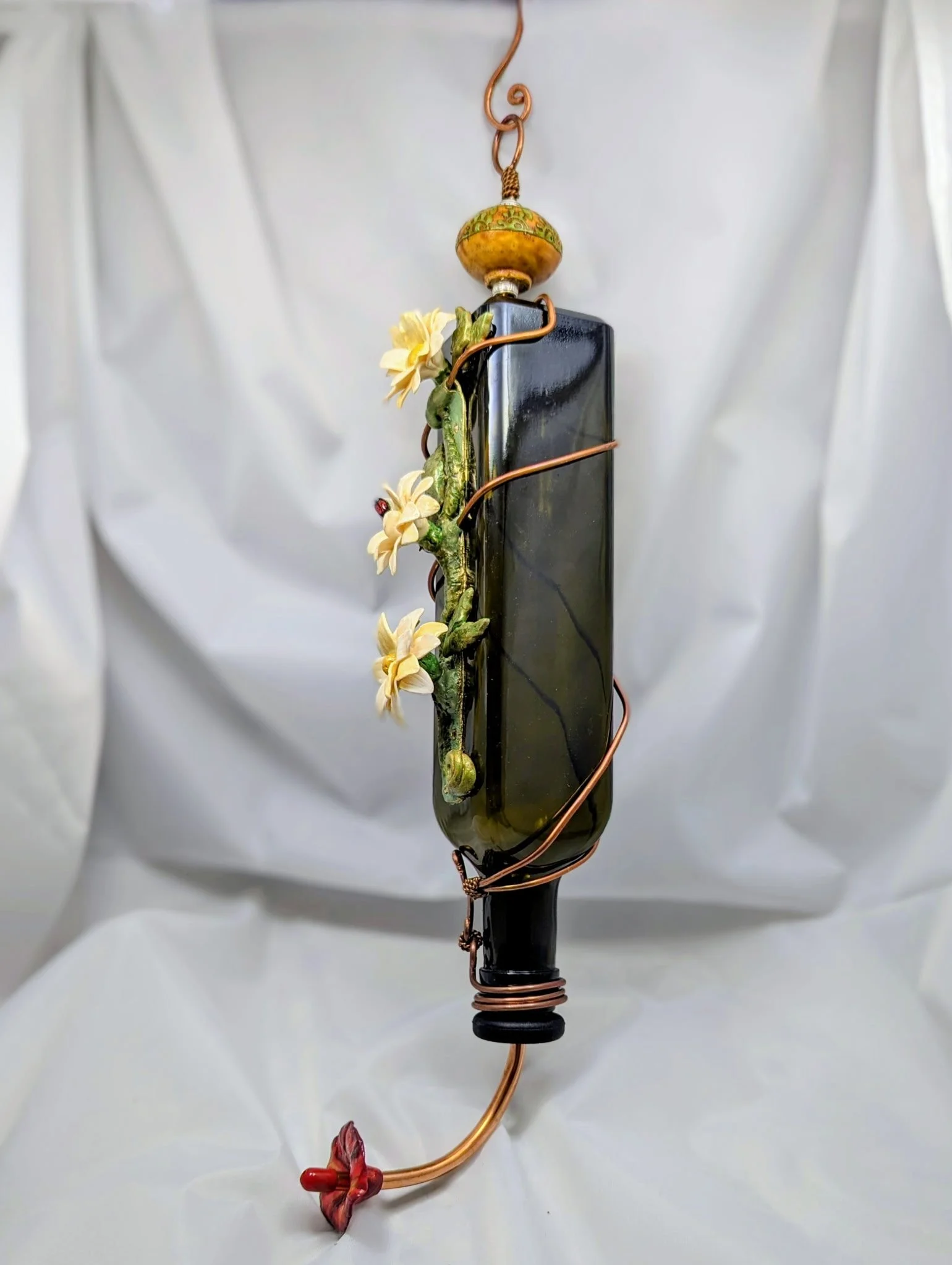 Handcrafted decorative hummingbird feeder by Art Illusions by Young featuring an orange and green ceramic knob, small white flowers, green leaves, and copper wire accents around a green glass bottle.