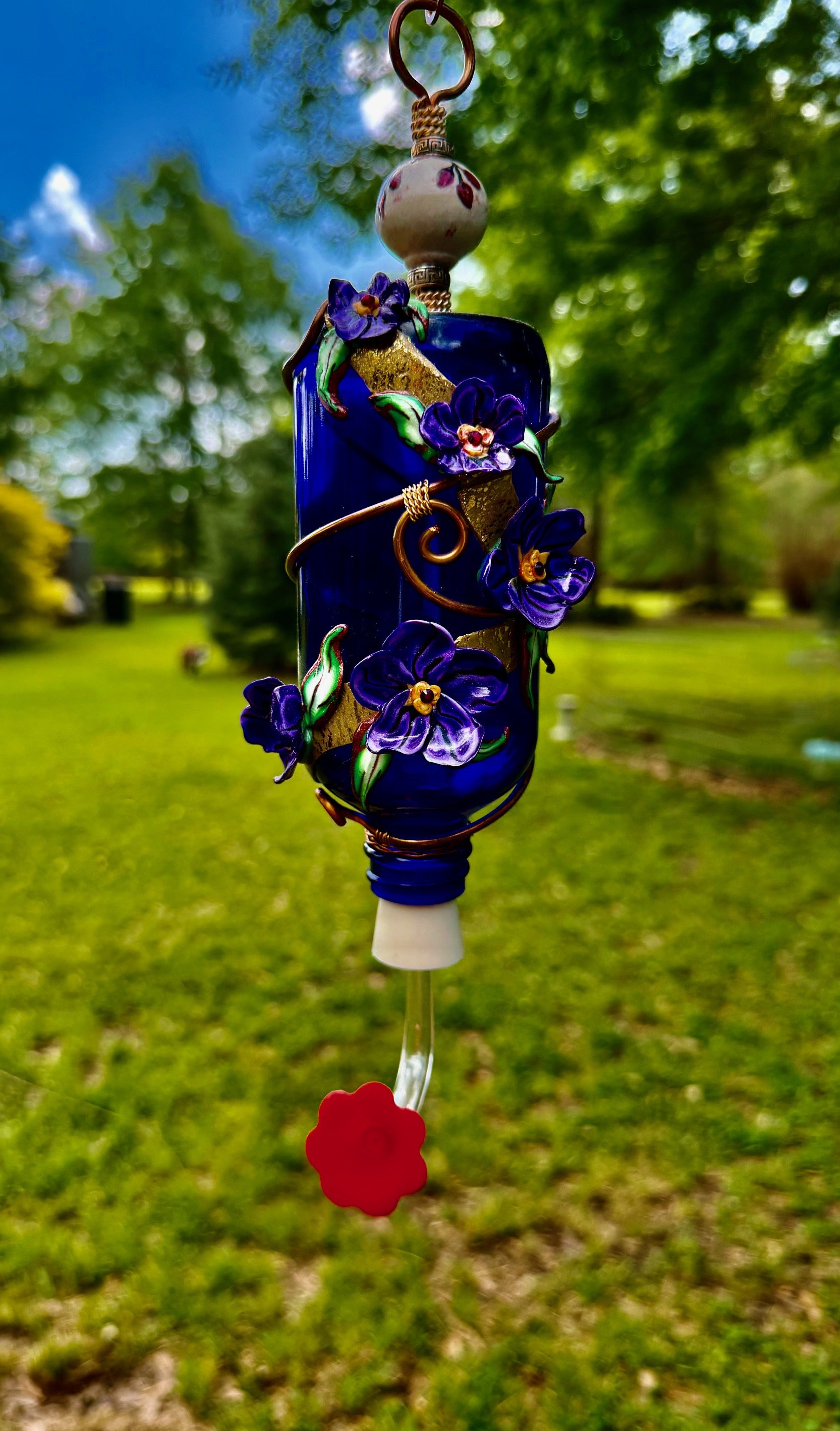 Colorful handcrafted hummingbird feeder by Art Illusions by Young hanging outdoors in a garden setting with green trees and blue sky in the background.