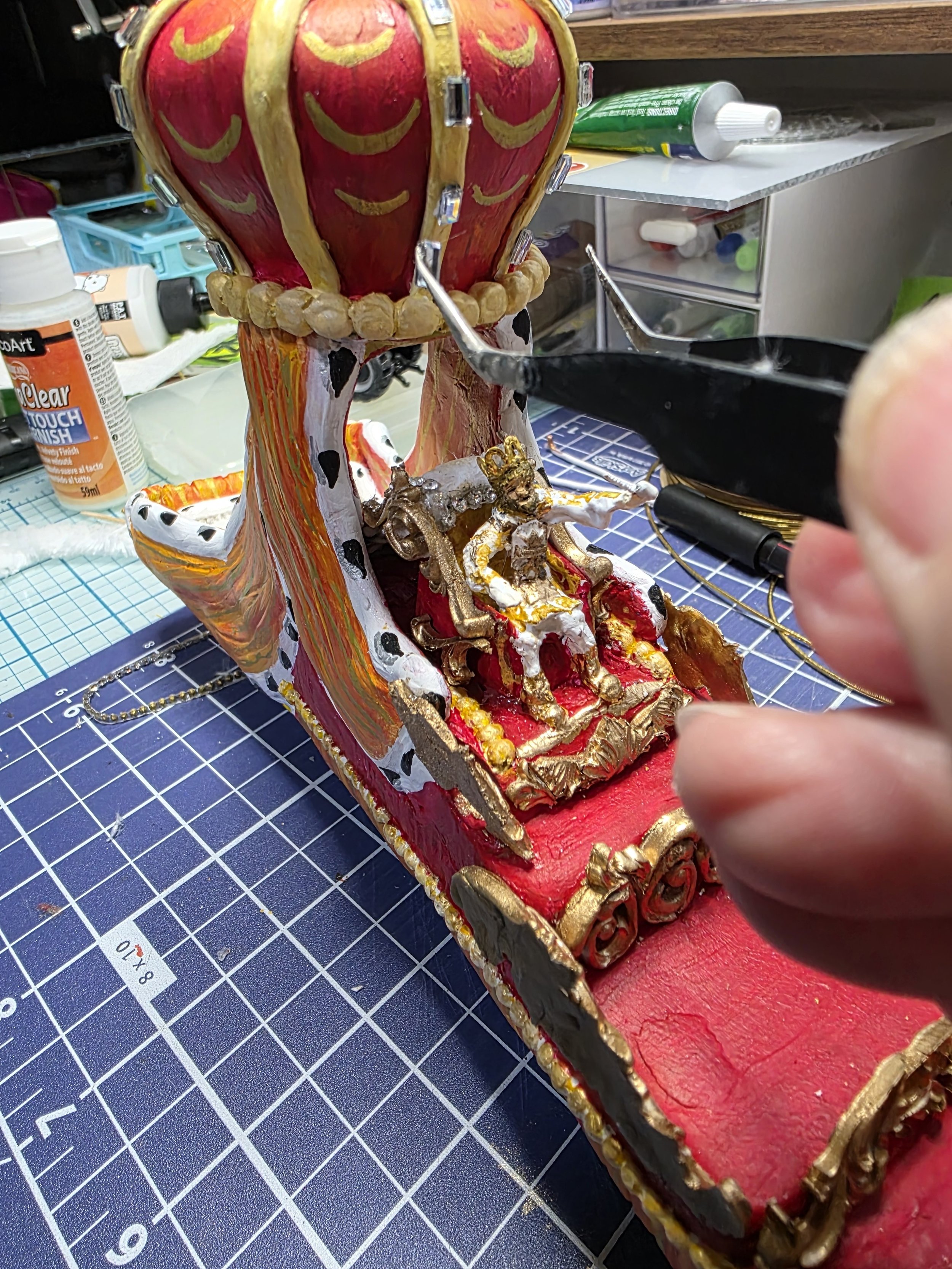 Close-up of hands assembling the ornate red and gold micro mirror sections of the King Rex parade float. Intricate handcrafting process of the mixed media diorama by Art Illusions by Young.