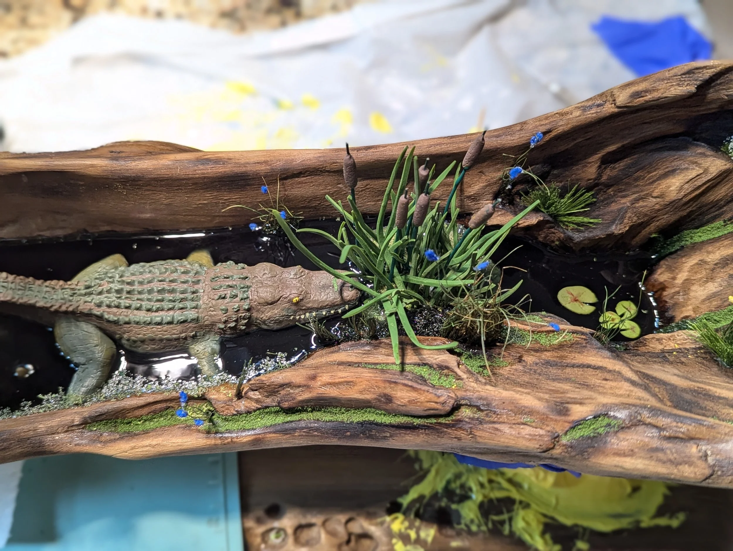 Handcrafted micro swamp diorama by Art Illusions by Young featuring a small sculpted alligator resting among green plants, cattails, blue flowers, and reflective black epoxy water on a natural driftwood base. Realistic Louisiana bayou wetland scene f