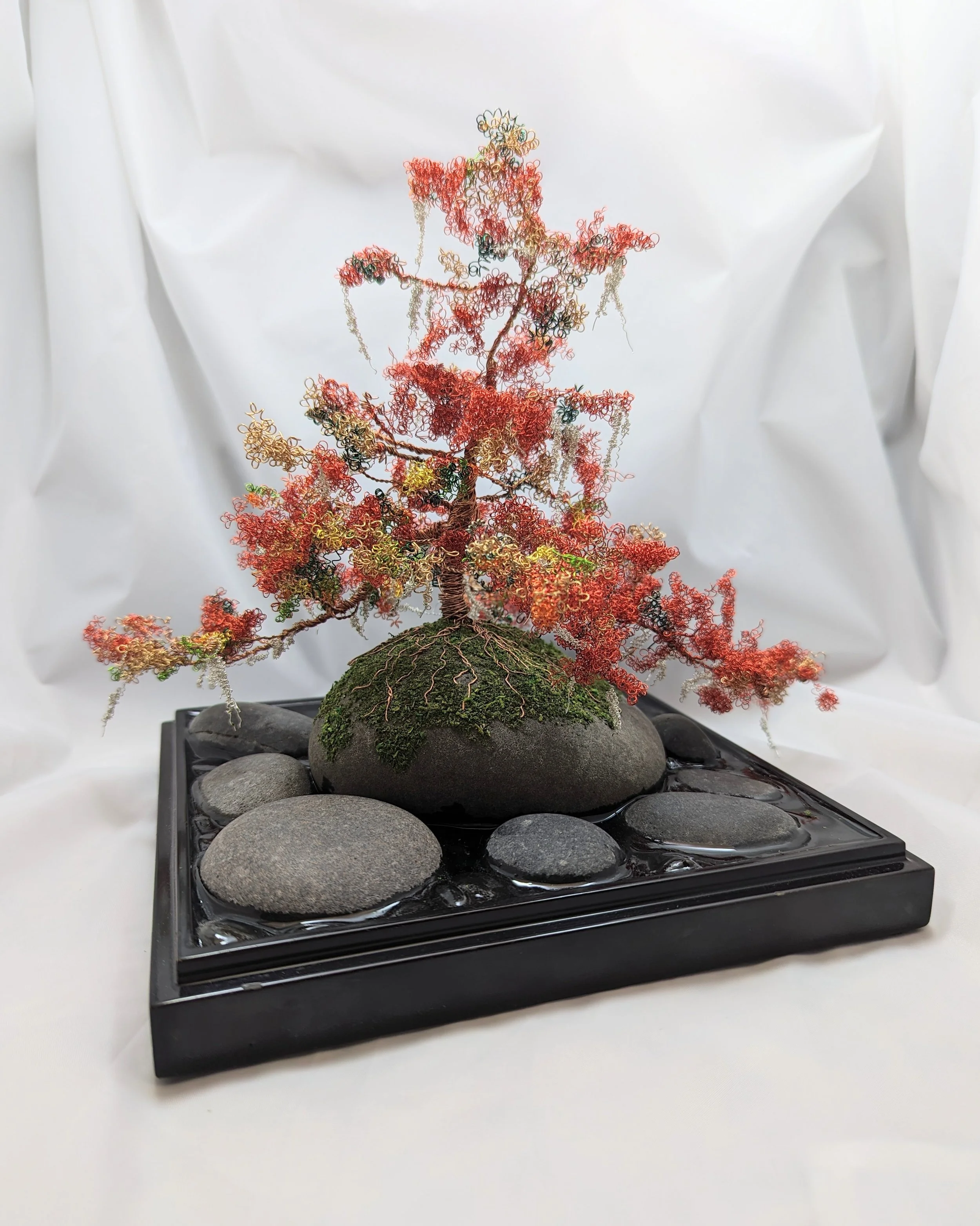 Flame of Autumn in Copper Wire bonsai diorama by Art Illusions by Young. Hand-woven red and green copper wire tree with autumn leaves on black stone base with river stones, moss, and black epoxy water pond. 110 hours work.