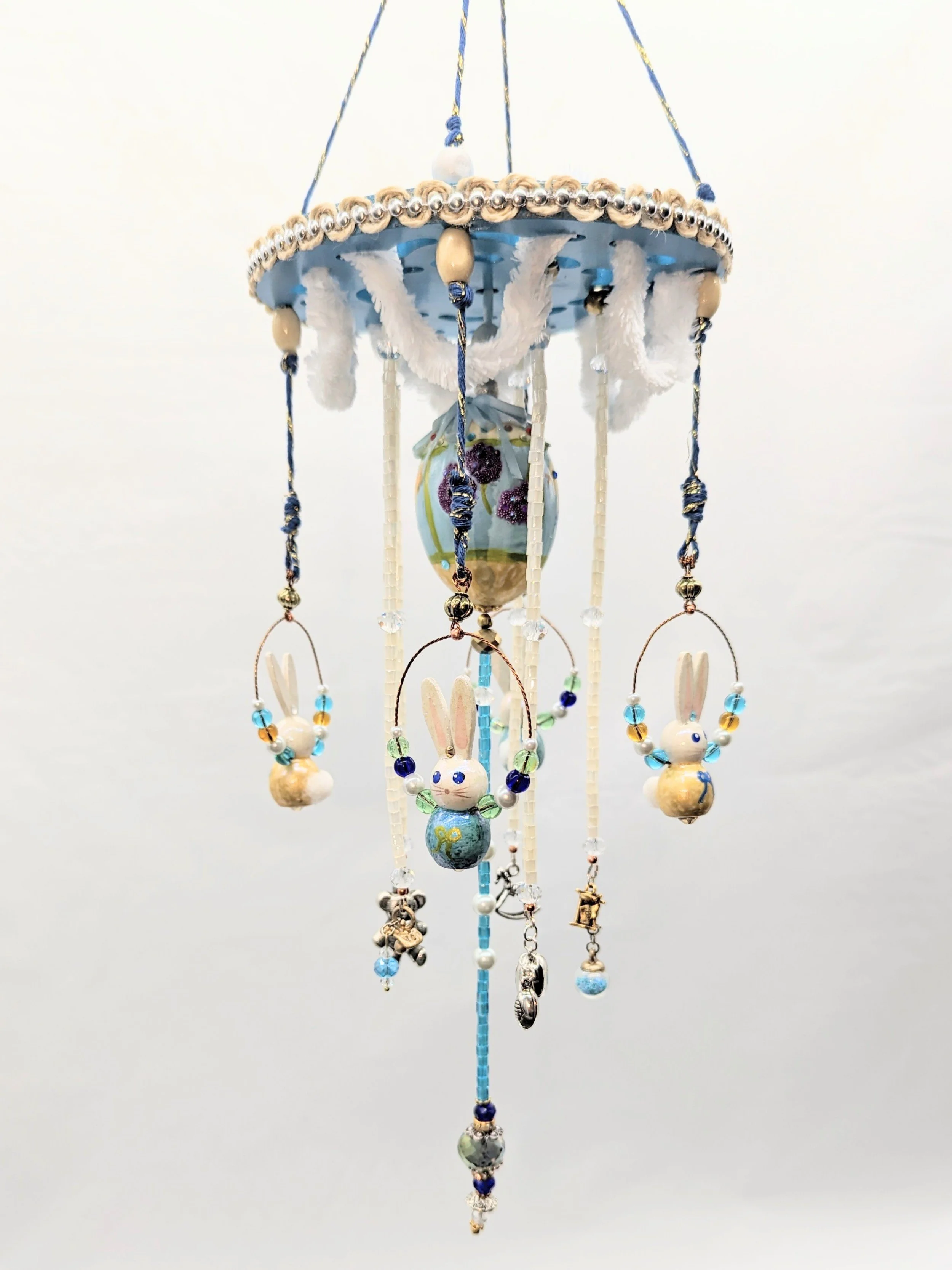 Full hanging view of the Baby Mobile – Cherished Nursery Keepsake by Art Illusions by Young. Measuring approximately 6” wide by 12” long, the mobile features the painted blue wooden disc with scalloped white fluffy cording and golden accents, four ha