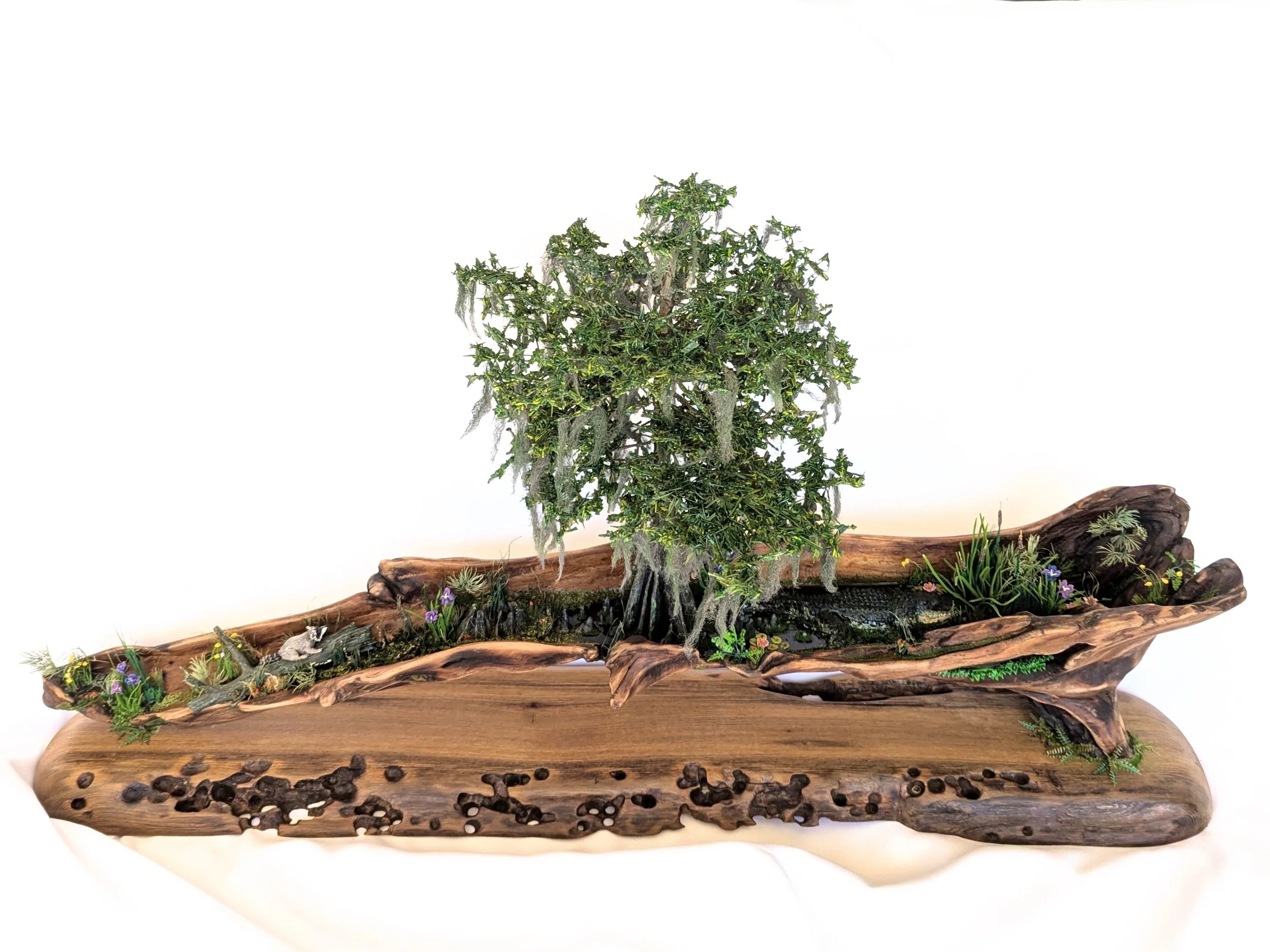 Miniature cypress tree on hollowed cypress and cypress driftwood base with subtle water accents, sculpted details, and foliage against white fabric. Part of the Micro Swamp / Bonsai collection by Art Illusions by Young.