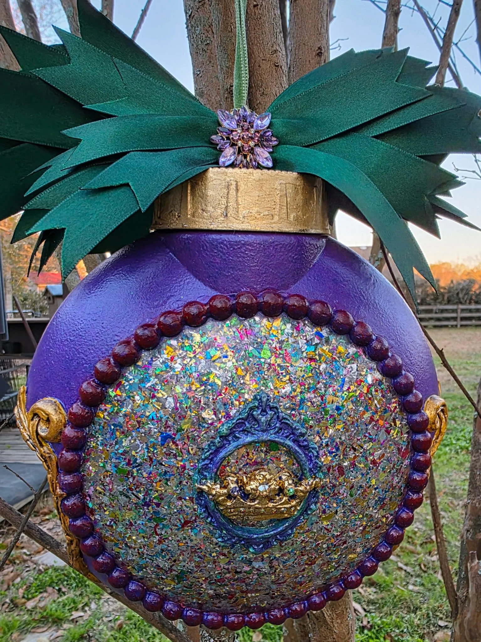 Close-up detail of the queen's crown on the reverse side of the Dual Crown Royalty Ornament by Art Illusions by Young. The lavish emerald-green ribbon bow and sparkling lavender-colored brooches accent the top. The multi-colored encased foil dome rev