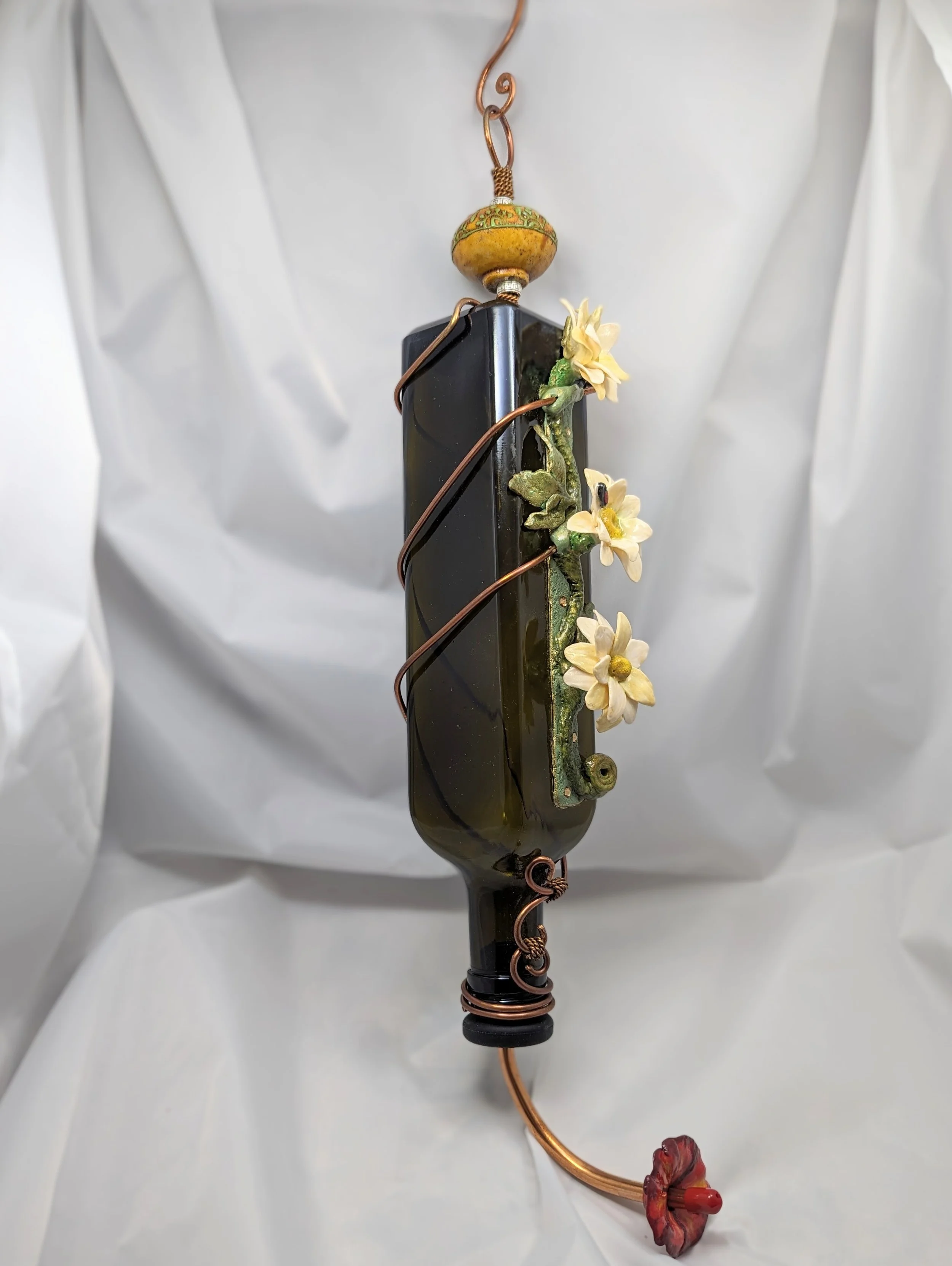 Left-angled view of the White Daffodil Elegance Hummingbird Feeder by Art Illusions by Young. Intricate copper wire is shaped into artistic vines and loops around the dark glass bottle, complemented by hand-sculpted white daffodils in polymer clay. T