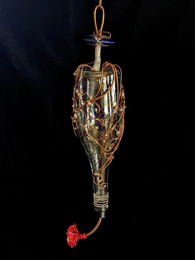 Right side view on black background of the Copper Spiral Grace Hummingbird Feeder by Art Illusions by Young. The copper wire is carefully shaped into flowing spirals and tendrils around the clear glass bottle, accented with a subtle hand-sculpted pol