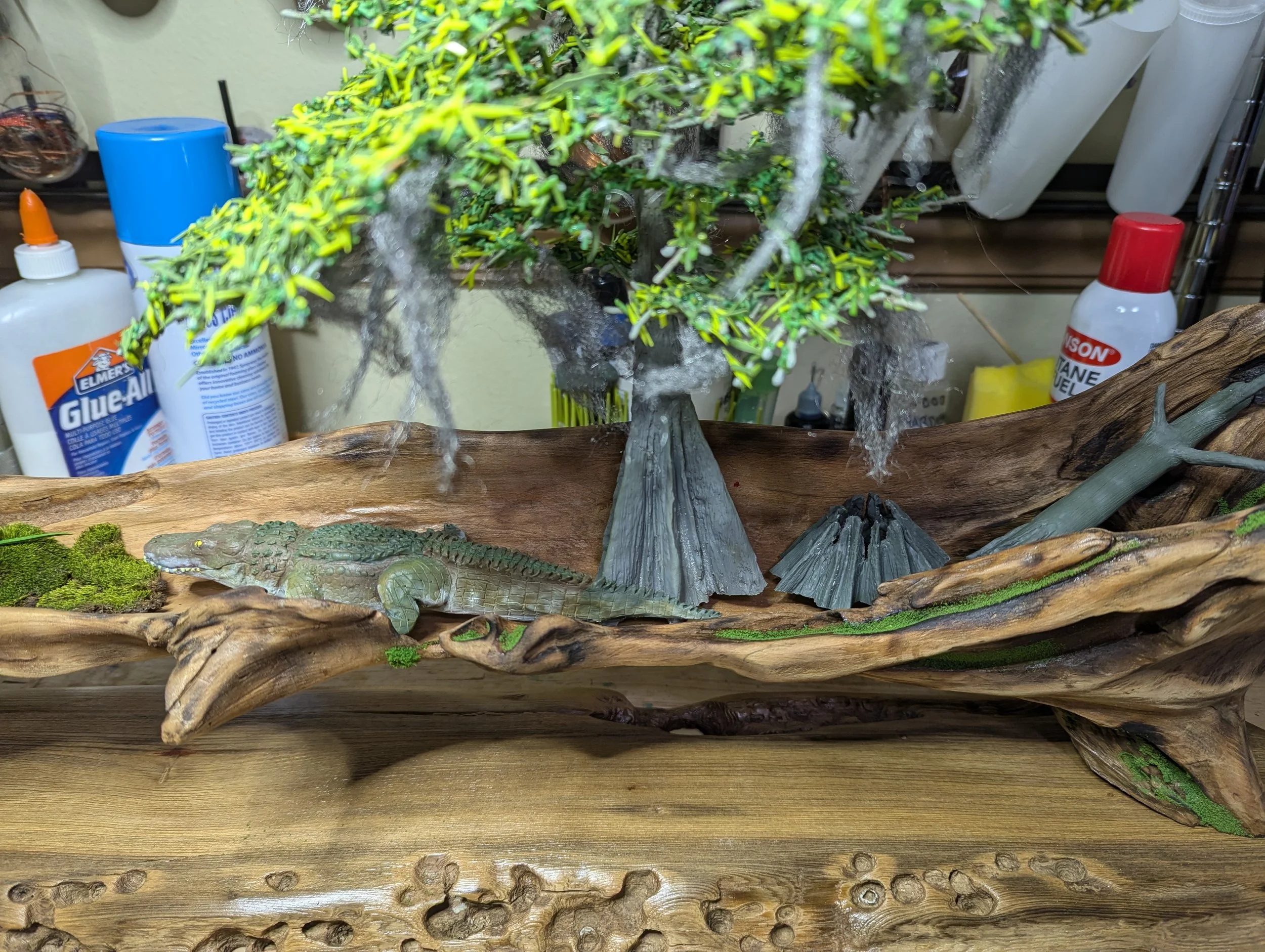 Pre-placement of sculptured alligator, cypress tree and stumps onto the hollowed cypress. Detailed micro swamp landscape on driftwood featuring a winding water channel, assorted grasses, small purple and white wildflowers, and textured soil layers. R