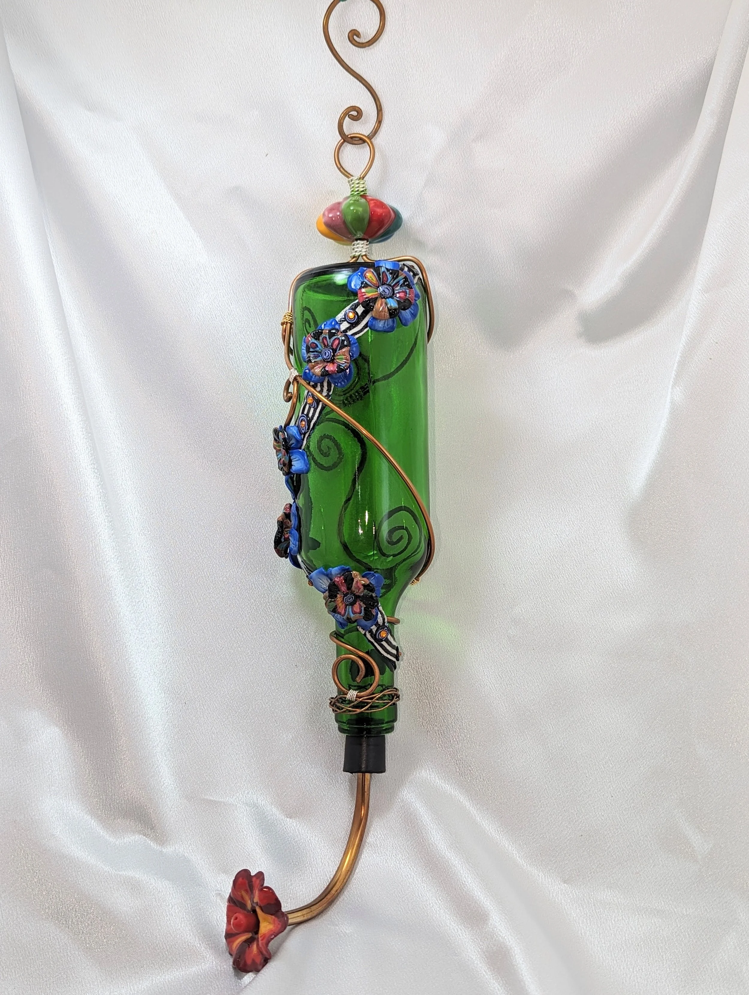 Green glass hummingbird feeder by Art Illusions by Young featuring colorful floral decorations and copper wire accents, hanging against a white satin fabric background.