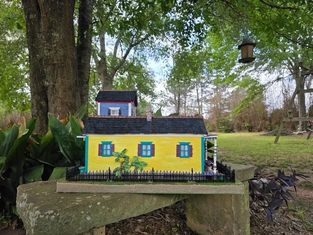 Left-side view of the handcrafted Triple New Orleans Shotgun House Row diorama by Art Illusions by Young. This angle highlights the sunny yellow shotgun house with vibrant teal and purple accents, detailed purple shutters, a ripe banana tree, ornate 