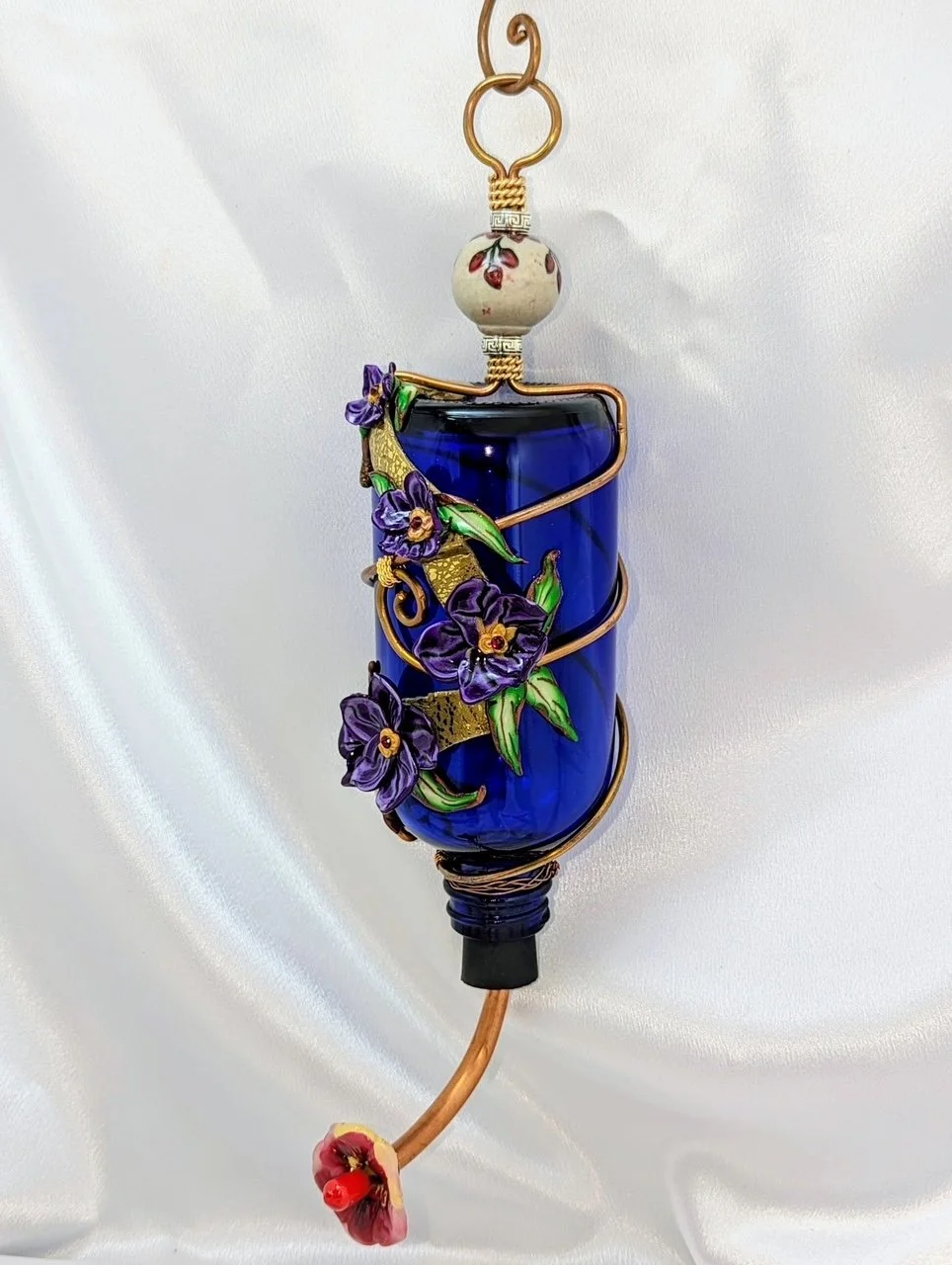 Right side frontal view detail of the Hummingbird Feeder Purple Pansy Elegance by Art Illusions by Young. The copper wire is wrapped and twisted into artistic patterns around the cobalt blue bottle, complemented by delicate hand-sculpted polymer clay