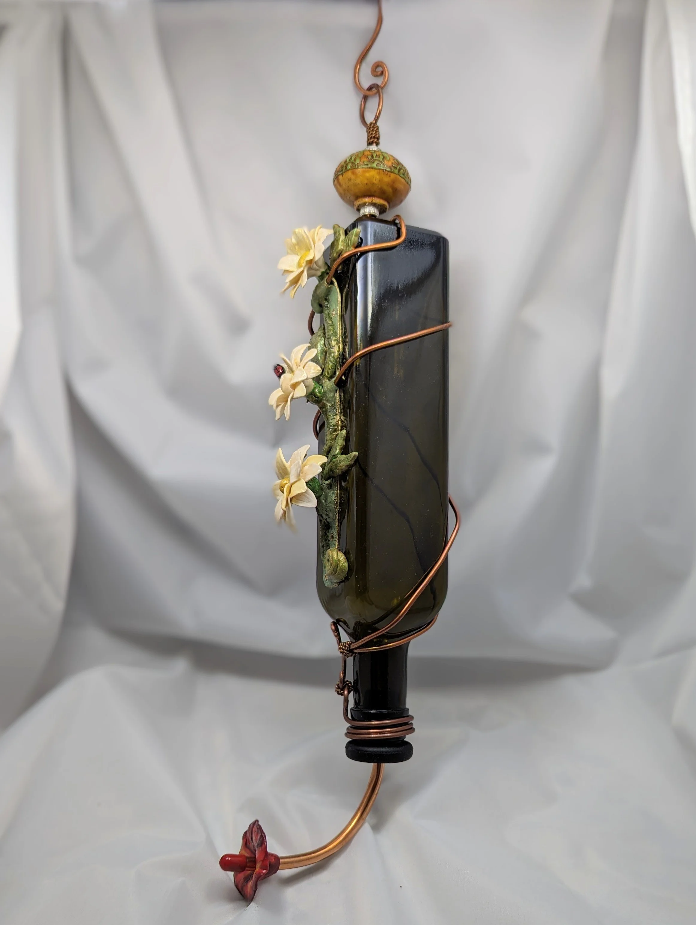 Right-angled view of the White Daffodil Elegance Hummingbird Feeder by Art Illusions by Young. The recycled dark green bottle features elegant copper wire wrapping and hand-sculpted white daffodils, finished with a secure ceramic knob and decorative 