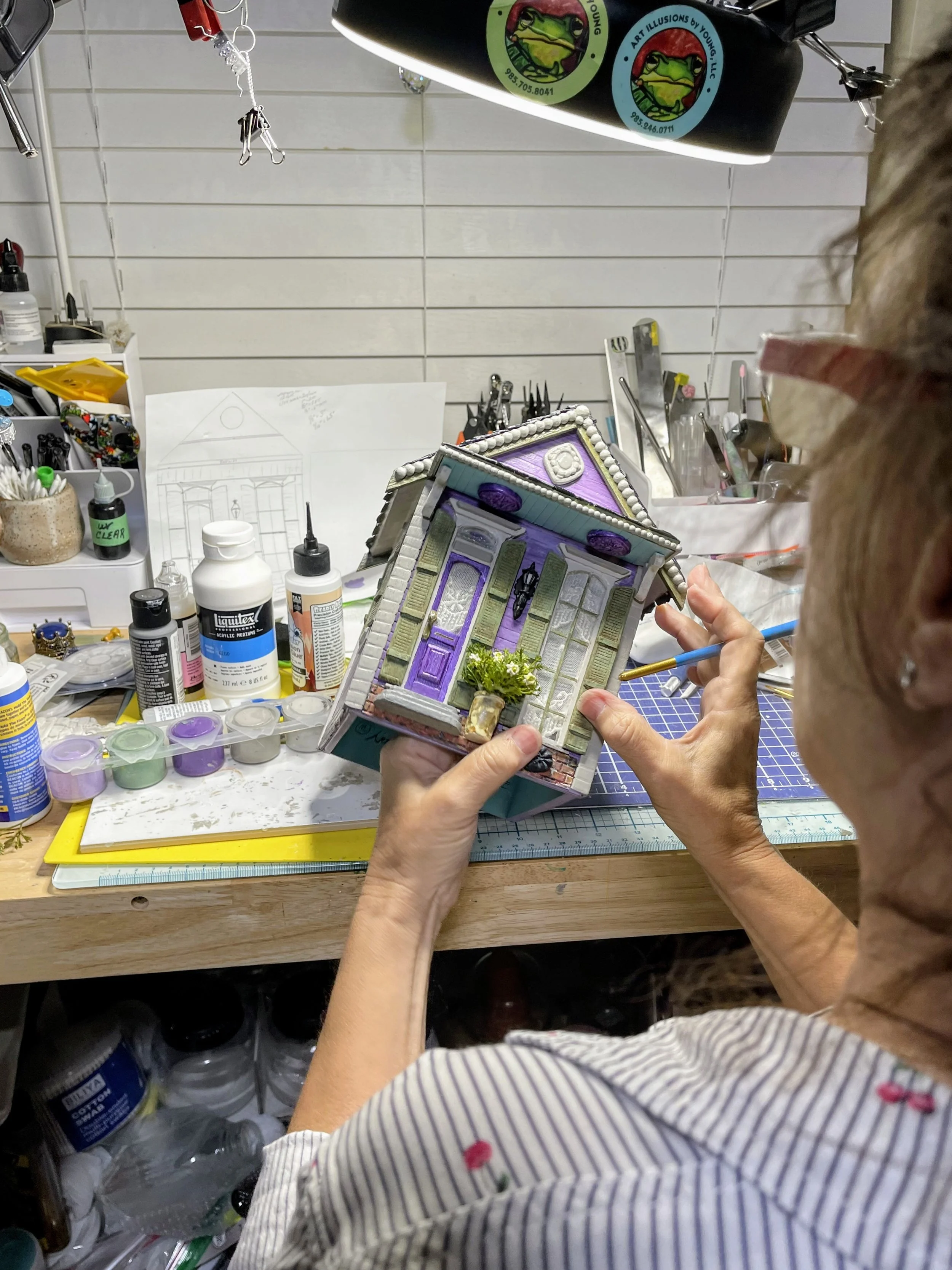 Artist Sandra Young holding the Purple Reign Shotgun House diorama to create detailing. When complete, the lavender shotgun house with black wrought iron fence, detailed yard, and functional tissue box holder is a one-of-a-kind mixed media functional