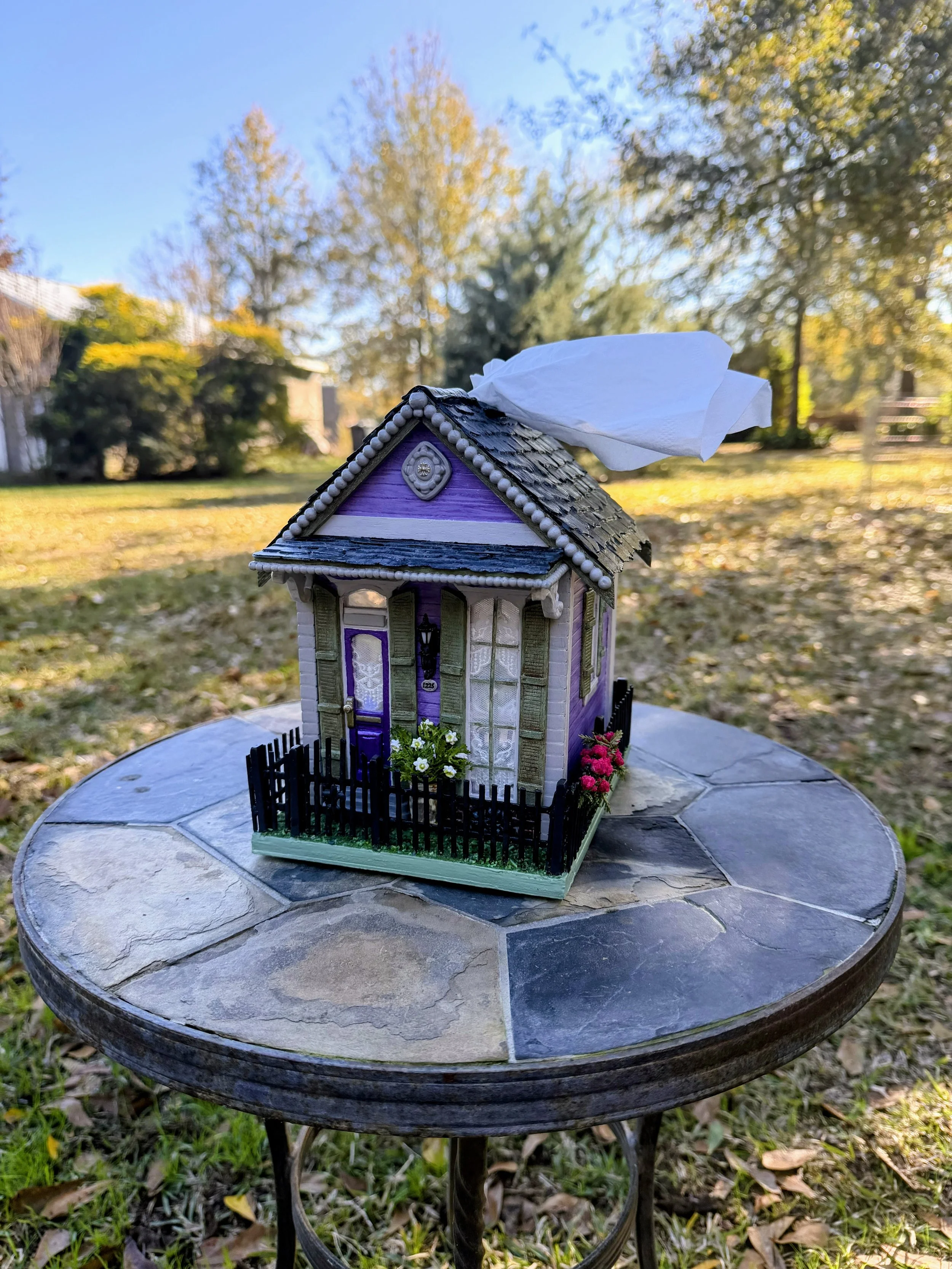 Angled view of the Purple Reign Shotgun House miniature diorama by Art Illusions by Young. Features a welcoming front porch with lantern, clay address plaque, potted clay gardenia flowers, and detailed wooden black "wrought iron" fence. Hand-sculpted