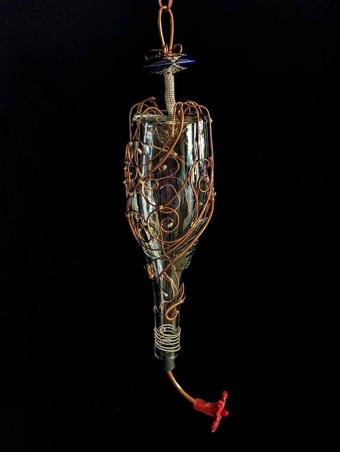 Left side view of the Copper Spiral Grace Hummingbird Feeder by Art Illusions by Young. The recycled clear glass wine bottle is adorned with elegant copper wire spirals and a minimal floral accent, demonstrating the meticulous craftsmanship invested 
