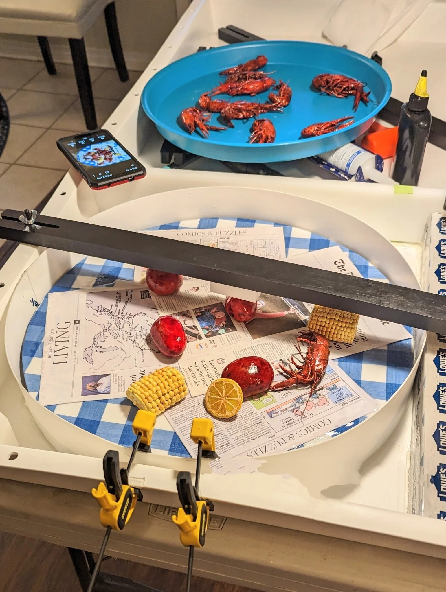 Studio assembly view of the Crawfish Boil Epoxy Wall Art by Art Illusions by Young. Sections of the full boil spread — taxidermy crawfish, hand-sculpted corn, lemons, and printed newspaper — is arranged on the work surface with tools visible, illustr