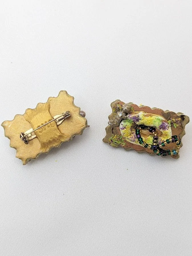 Two Mardi Gras King Cake Baby magnets/pins by Art Illusions by Young shown back and front on a clean white background. The hand-sculpted clay babies feature gold-foiled fronted clay and colorful bead accents in purple, green, and gold. These playful 