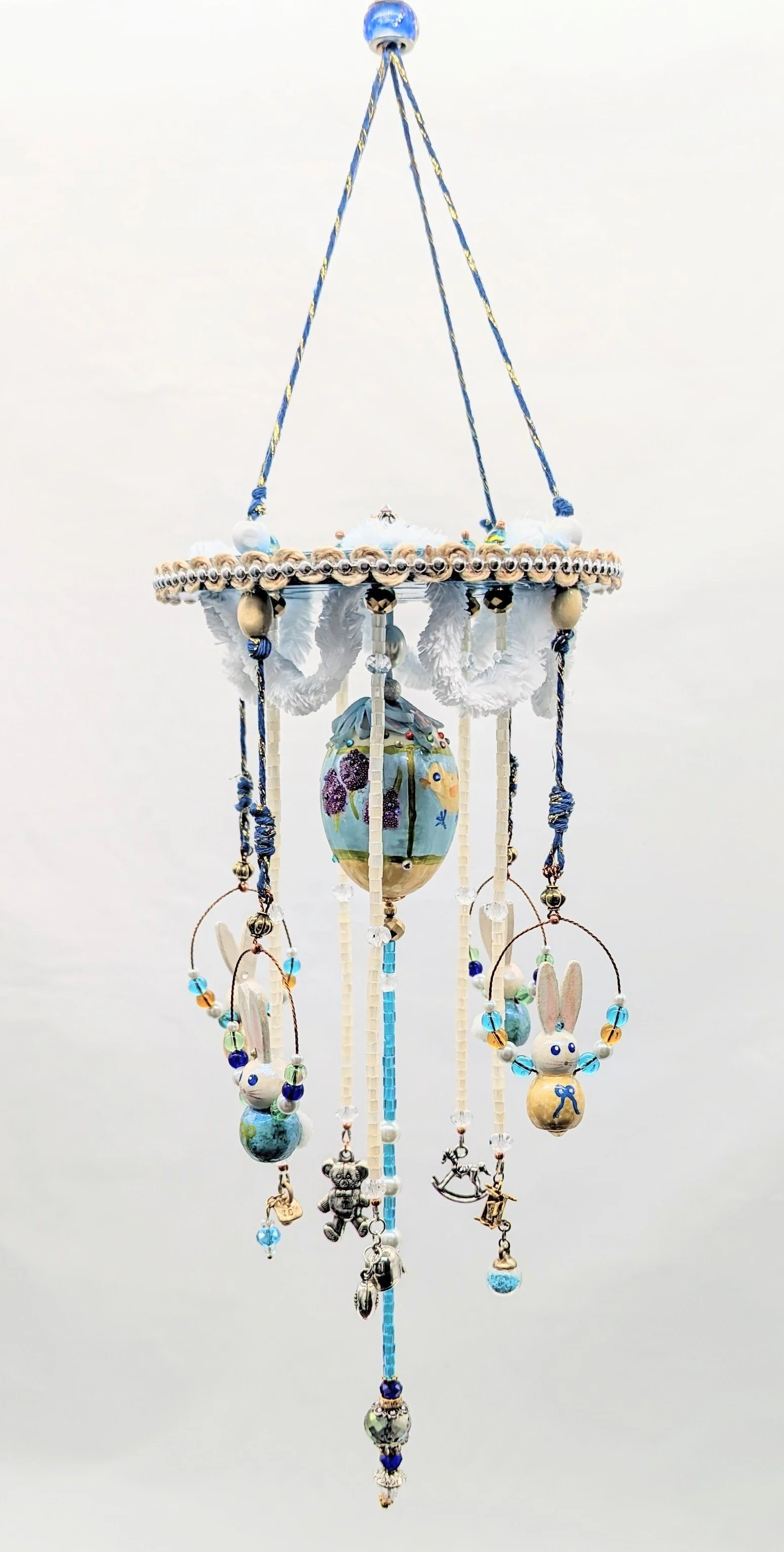 Studio view of the Baby Mobile – Cherished Nursery Keepsake by Art Illusions by Young. The painted blue wooden disc with scalloped white fluffy cording and golden accents is shown with the hand-painted wooden bunnies embellished with epoxy, crystals,
