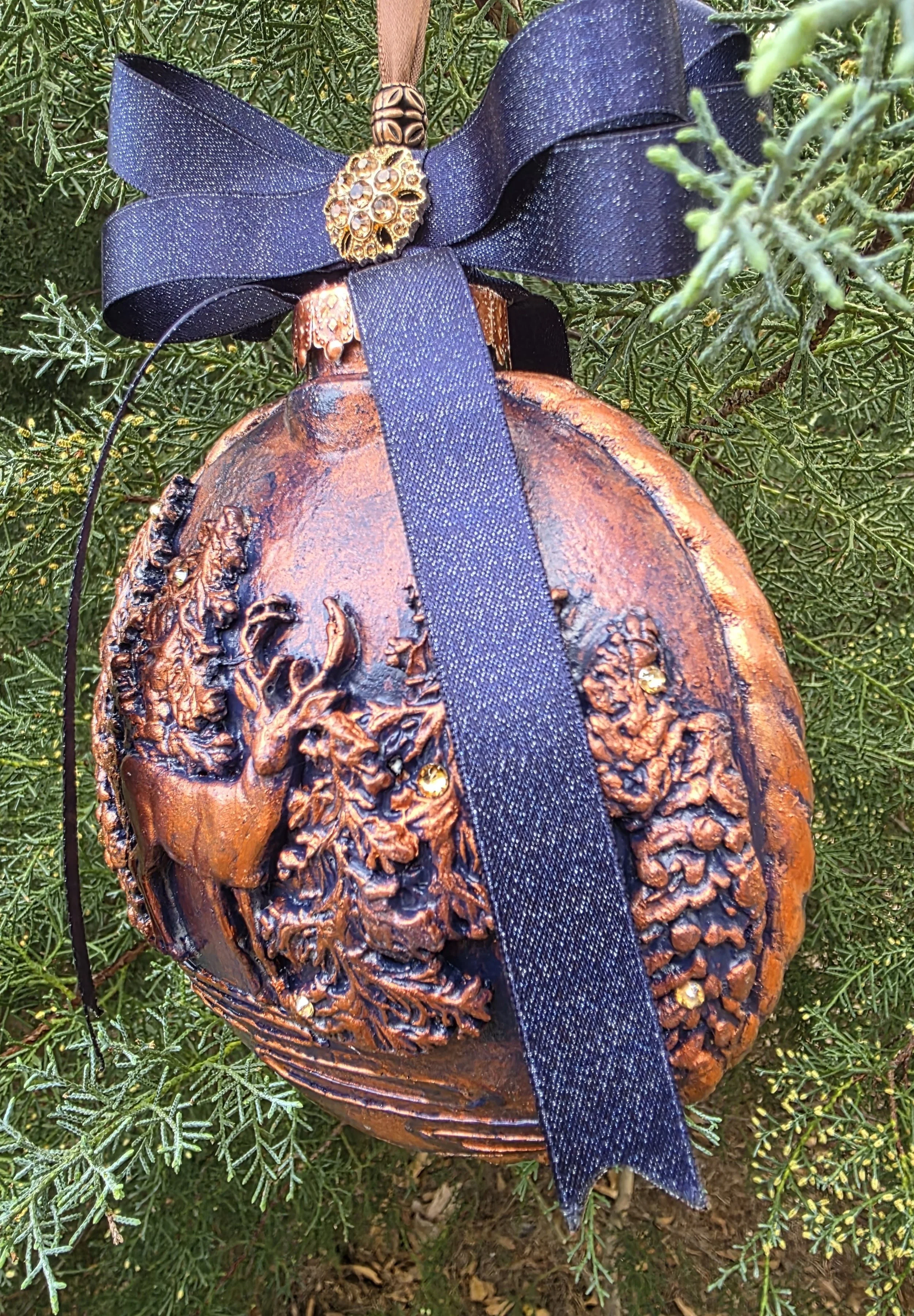Angled view of the Woodland Majesty Ornament by Art Illusions by Young, shown with a ruler for scale. The hand-sculpted molded stag and surrounding pines and foliage display rich copper-bronze tones and textured relief details. A navy-blue ribbon bow