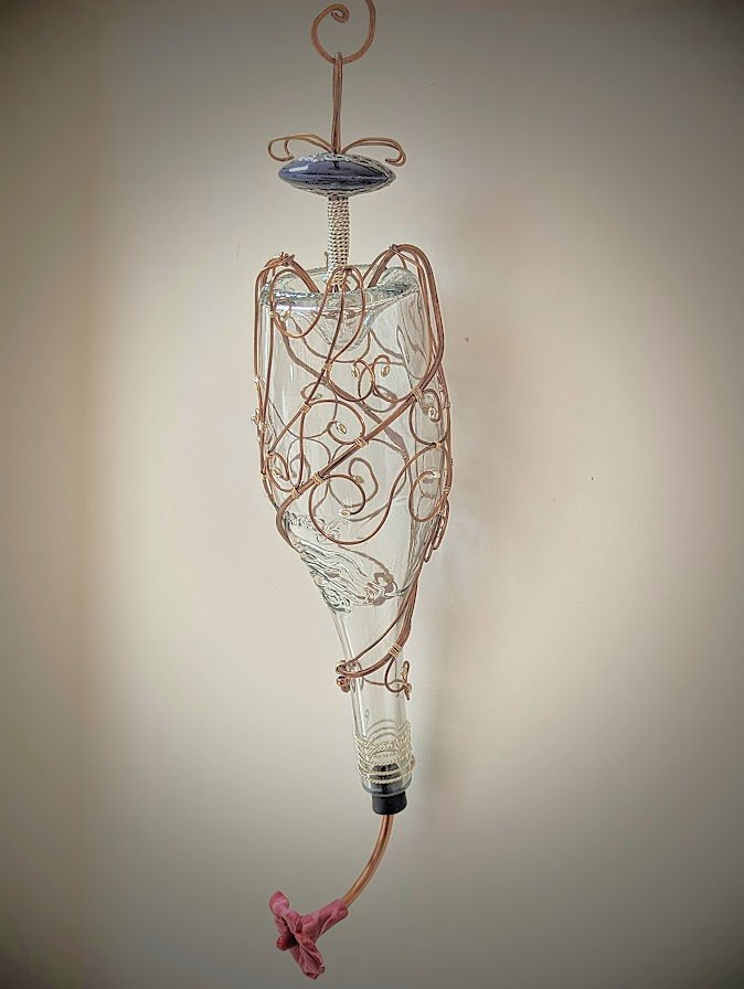 Intricate Copper Lace Hummingbird Feeder by Art Illusions by Young, Folsom, Louisiana. A recycled clear glass wine bottle is gracefully enveloped in hand-shaped copper wire twisted into delicate, lace-like organic patterns that spiral from base to ha