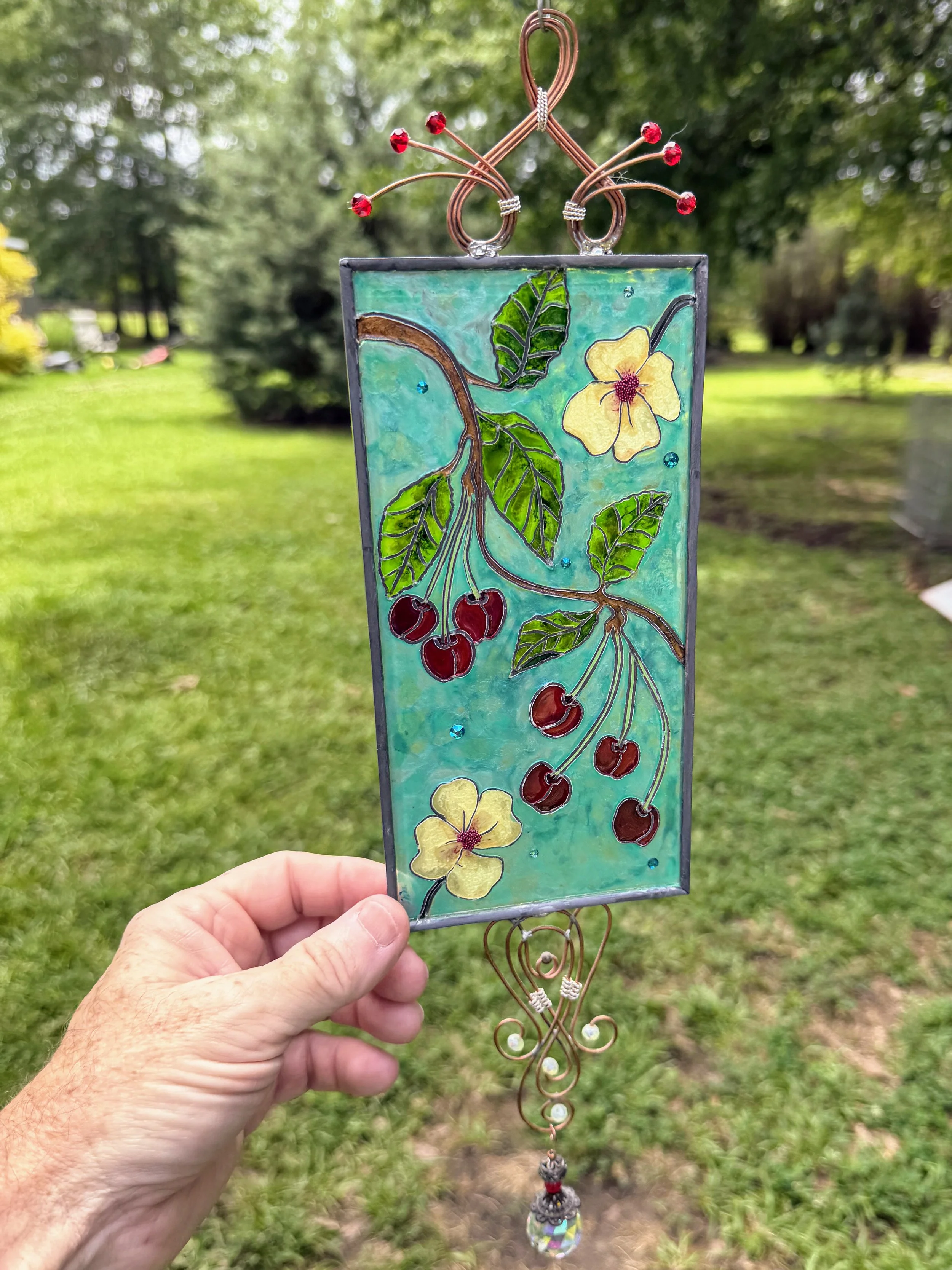 Cherry Jubilee by Art Illusions by Young. A joyful celebration of summer’s bounty bursts with life in this hand-painted and leaded Illusion Glass panel, shown here to emphasize its painterly quality. Clusters of ripe, glossy red cherries hang from de