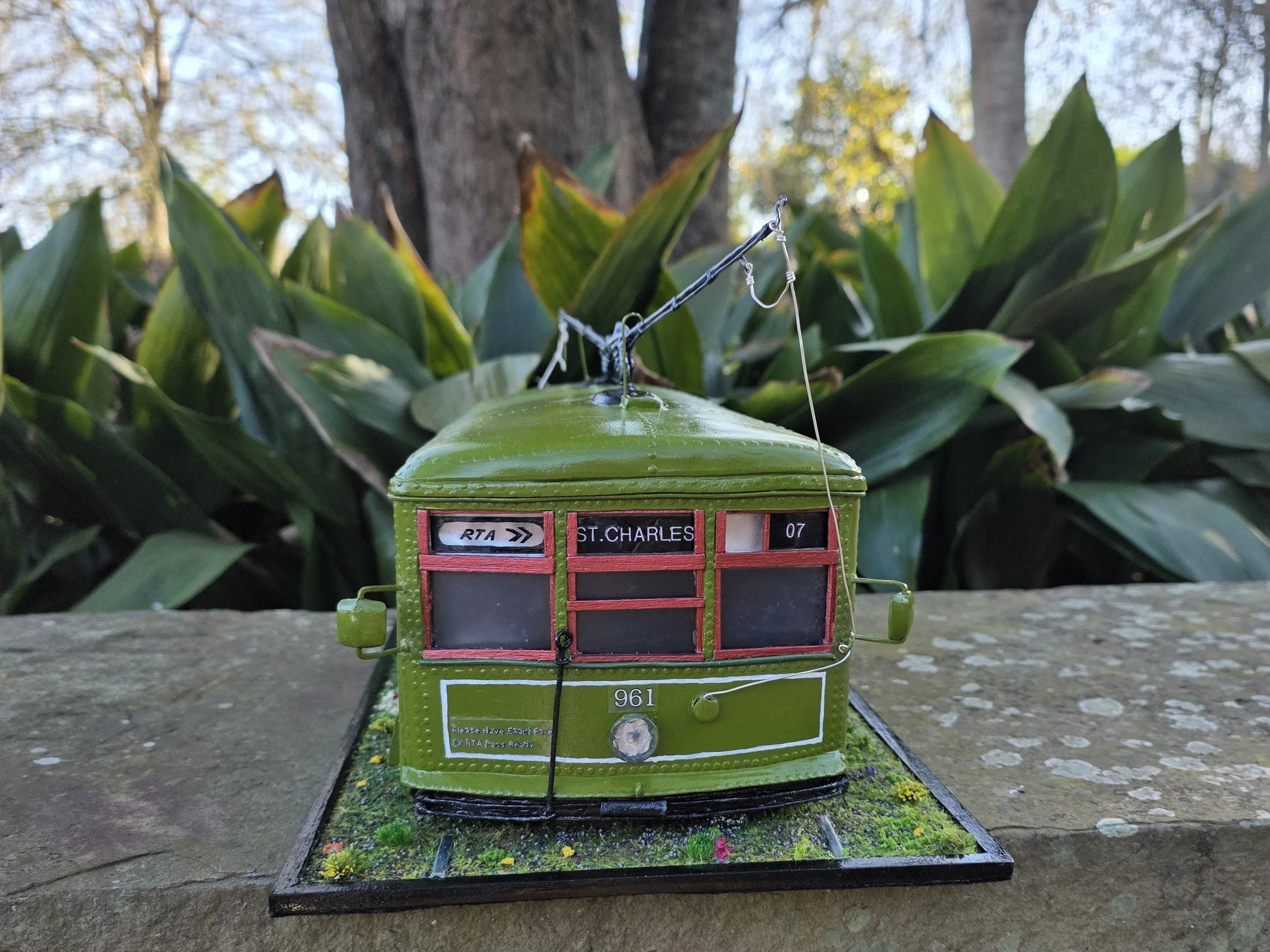Front three-quarter view of the hand-sculpted New Orleans streetcar tissue box cover featuring olive green body, red doors and windows, pantograph, and realistic details. Functional mixed media diorama by Art Illusions by Young in Folsom, Louisiana.